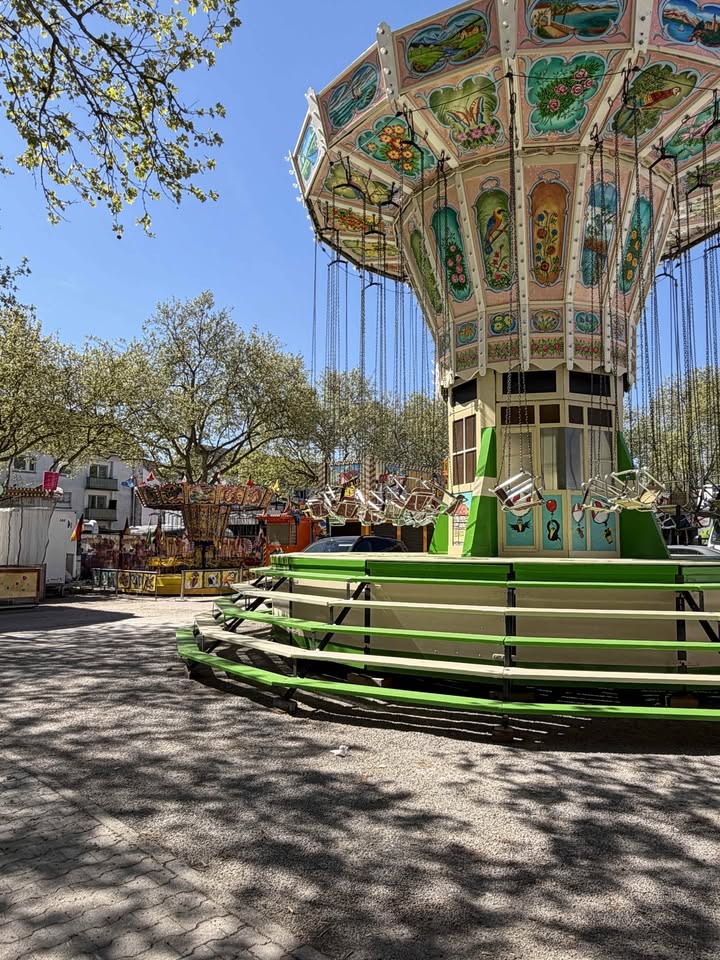 Dieser Alternativtext wurde KI-generiert: Ein farbenfrohes Karussell steht im Freien, umgeben von Bäumen und weiteren Attraktionen. Der Himmel ist blau und die Szene wirkt belebt.