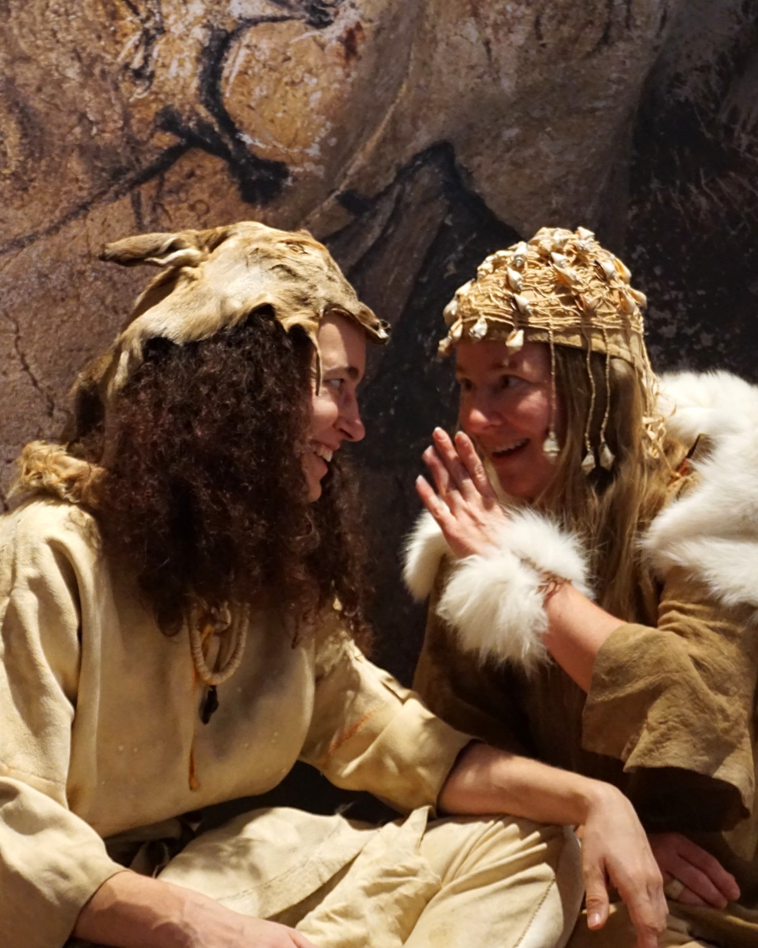 Dieser Alternativtext wurde KI-generiert: Zwei Personen tragen historische Kopfbedeckungen und Kleidung aus Naturmaterialien. Sie blicken einander lächelnd zu.