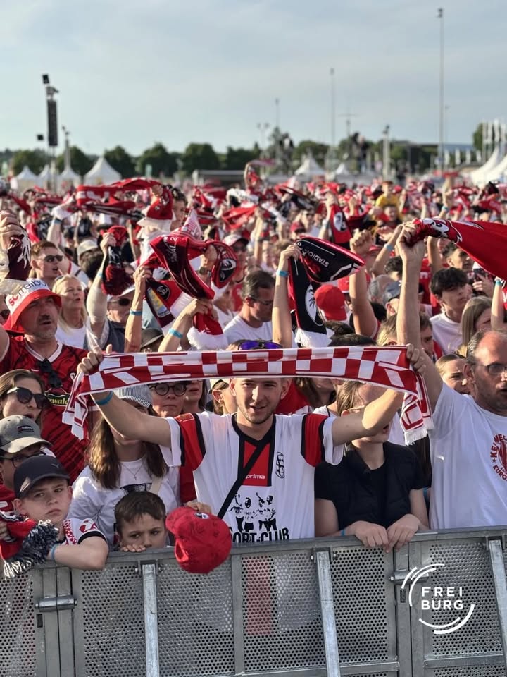 Menschenmenge vor einem Metallzaun, viele halten rot-weiße Schals hoch, einige tragen Trikots und Kopfbedeckungen, im Hintergrund Zelte und Bäume. (KI-Alternativtext)