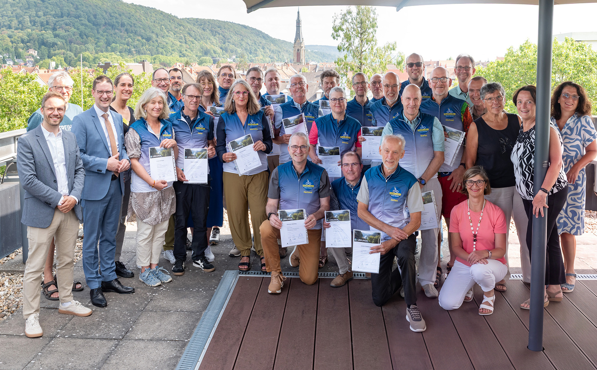 Eine große Gruppe von Personen posiert auf einer Holzterrasse vor einer Stadtlandschaft mit Kirchturm und bewaldetem Hügel. Viele der Teilnehmer tragen blaue Westen und halten weiße Urkunden in die Höhe. Im Vordergrund knien einige Personen, während der Rest der Gruppe im Hintergrund steht. (KI-Alternativtext)