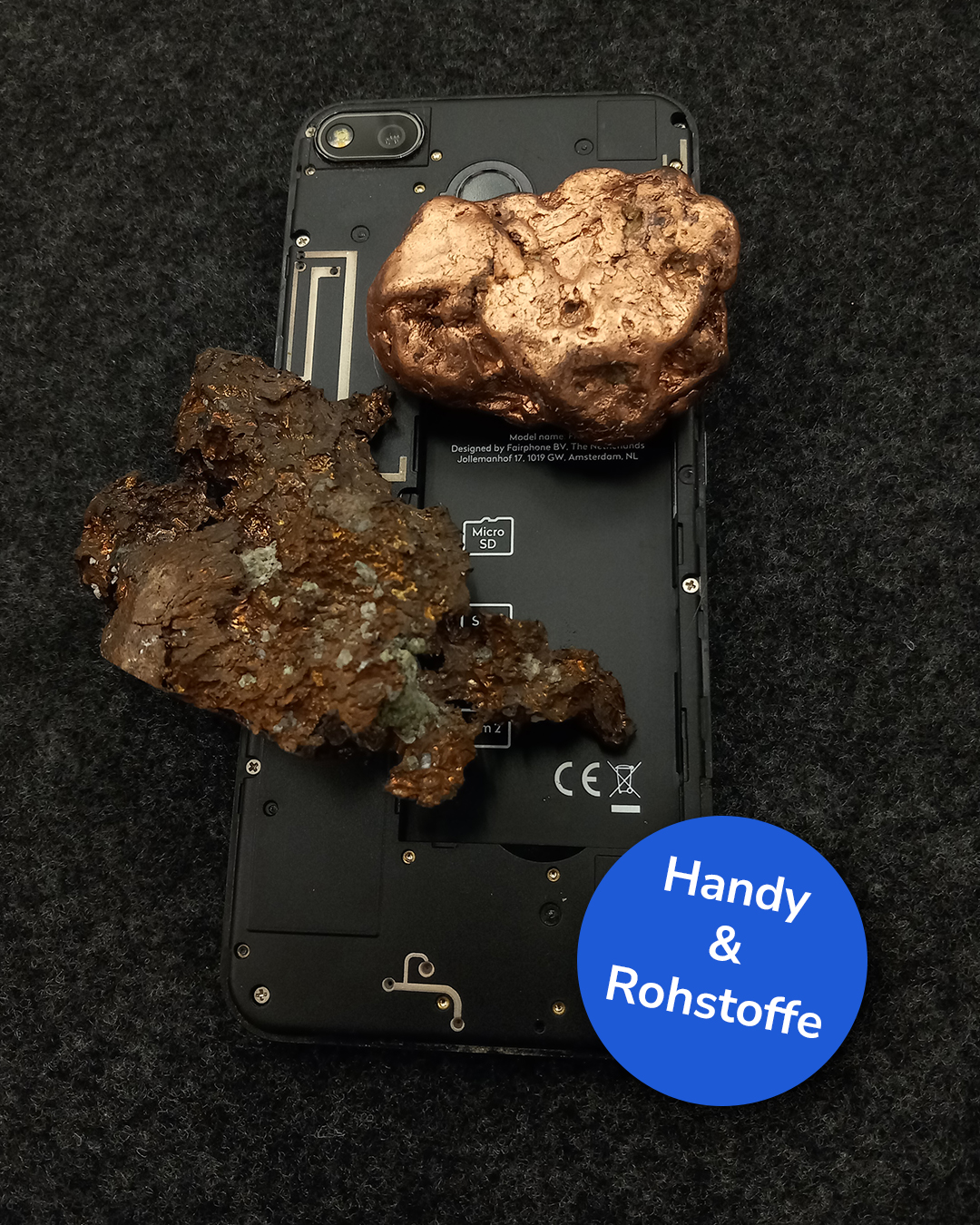Dieser Alternativtext wurde KI-generiert: Ein Mobiltelefon liegt auf einer dunklen Oberfläche neben mehreren rostfarbenen Mineralien. Das Bild ist farbig und zeigt auch Text und Symbole auf dem Telefon sowie Pfeile mit Beschriftungen.