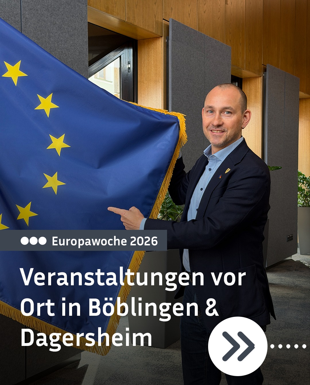 Dieser Alternativtext wurde KI-generiert: Eine Person steht vor einer Flagge mit gelben Sternen und einem Banner mit Text. Im Hintergrund sind graue Wandpaneele und ein Fenster zu sehen.
