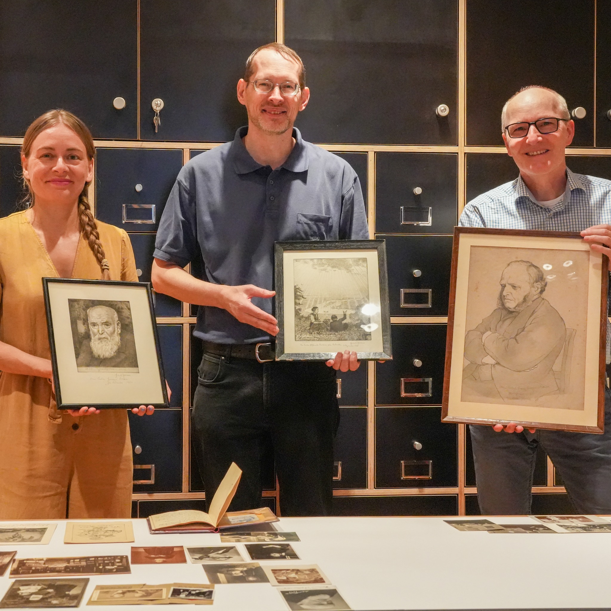 Drei Personen stehen vor dunkelblauen Schränken und halten jeweils ein gerahmtes historisches Bild oder eine Zeichnung. Auf dem weißen Tisch im Vordergrund sind weitere alte Fotos, Postkarten und ein offenes Buch ausgelegt. (KI-Alternativtext)