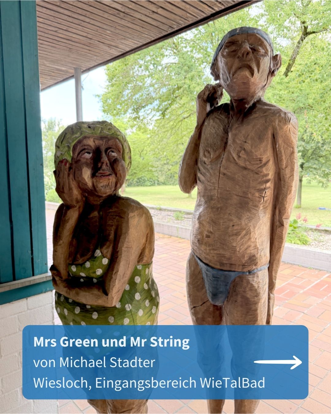 Zwei hölzerne Skulpturen älterer Personen stehen nebeneinander auf einer gepflasterten Fläche unter einem Dach. Die linke Figur trägt ein grünes Kleid mit weißen Punkten und eine Kopfbedeckung, die rechte Figur ist oberkörperfrei mit blauer Unterwäsche und Kopfbedeckung. Ein blauer Textkasten unten enthält die Aufschrift: „Mrs Green und Mr String von Michael Stadter Wiesloch, Eingangsbereich WieTalBad“ und einen Pfeil nach rechts. (KI-Alternativtext)