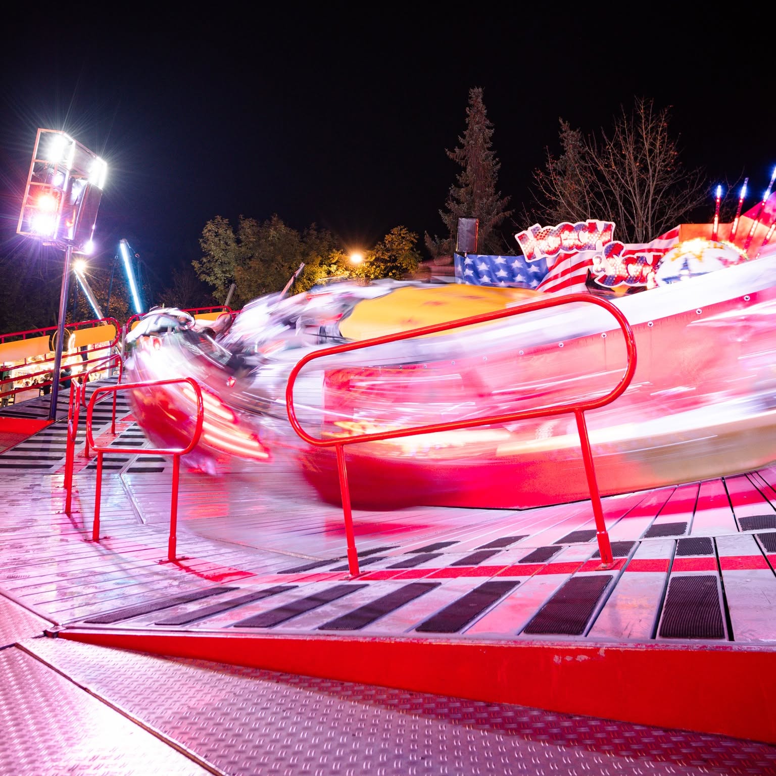 Dieser Alternativtext wurde KI-generiert: Eine beleuchtete Fahrgeschäftszene bei Nacht. Das rot-gelbe Fahrgeschäft ist in Bewegung, im Hintergrund sind weitere Lichter und Bäume zu sehen.