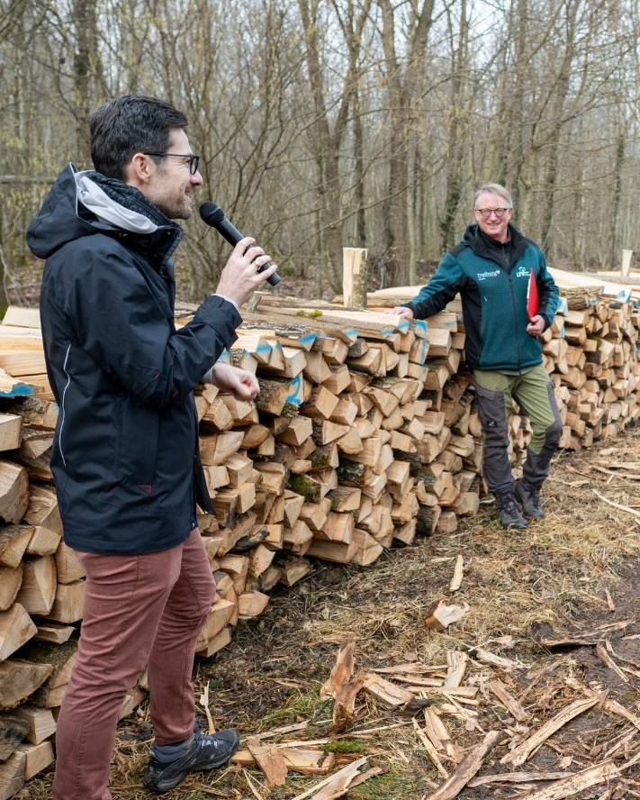 Dieser Alternativtext wurde KI-generiert: Zwei Personen stehen im Wald neben einem Holzstapel. Eine Person hält ein Mikrofon, die andere trägt eine rote mappe.