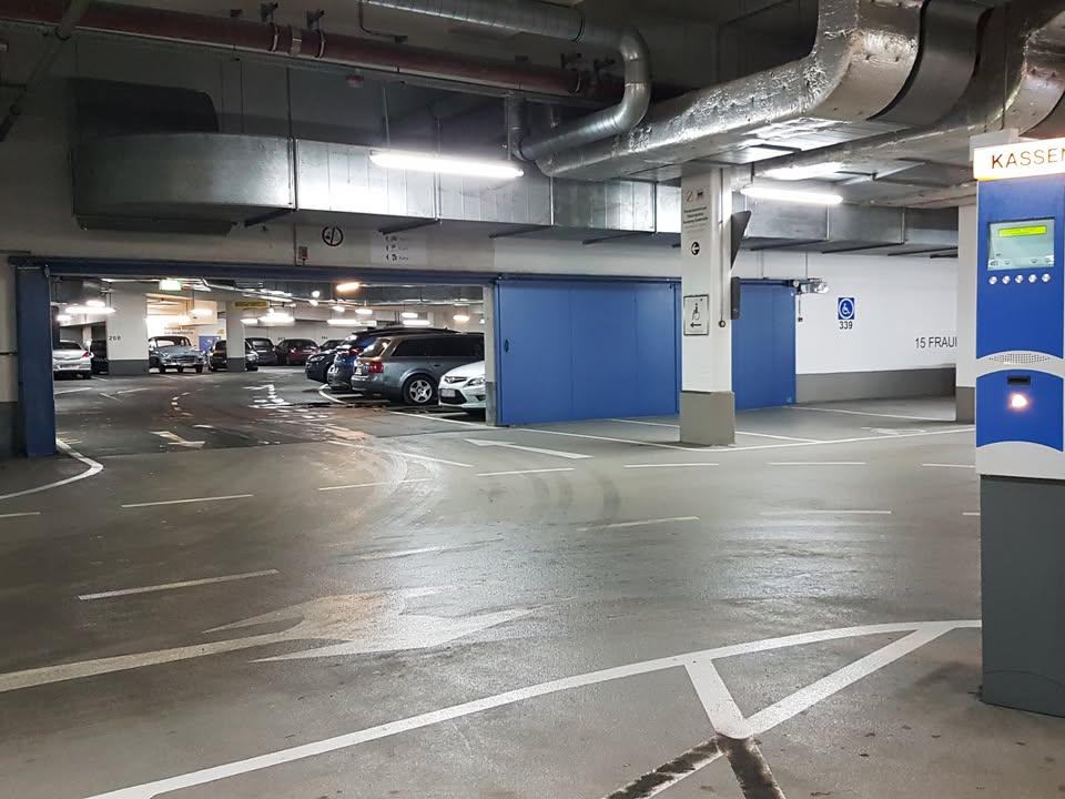 Dieser Alternativtext wurde KI-generiert: Ein Innenraum mit Fahrzeugen, markierten Parkplätzen und einer blauen Wand mit einem Kassenautomaten. Der Raum ist durch fluoreszierende Beleuchtung erhellt.