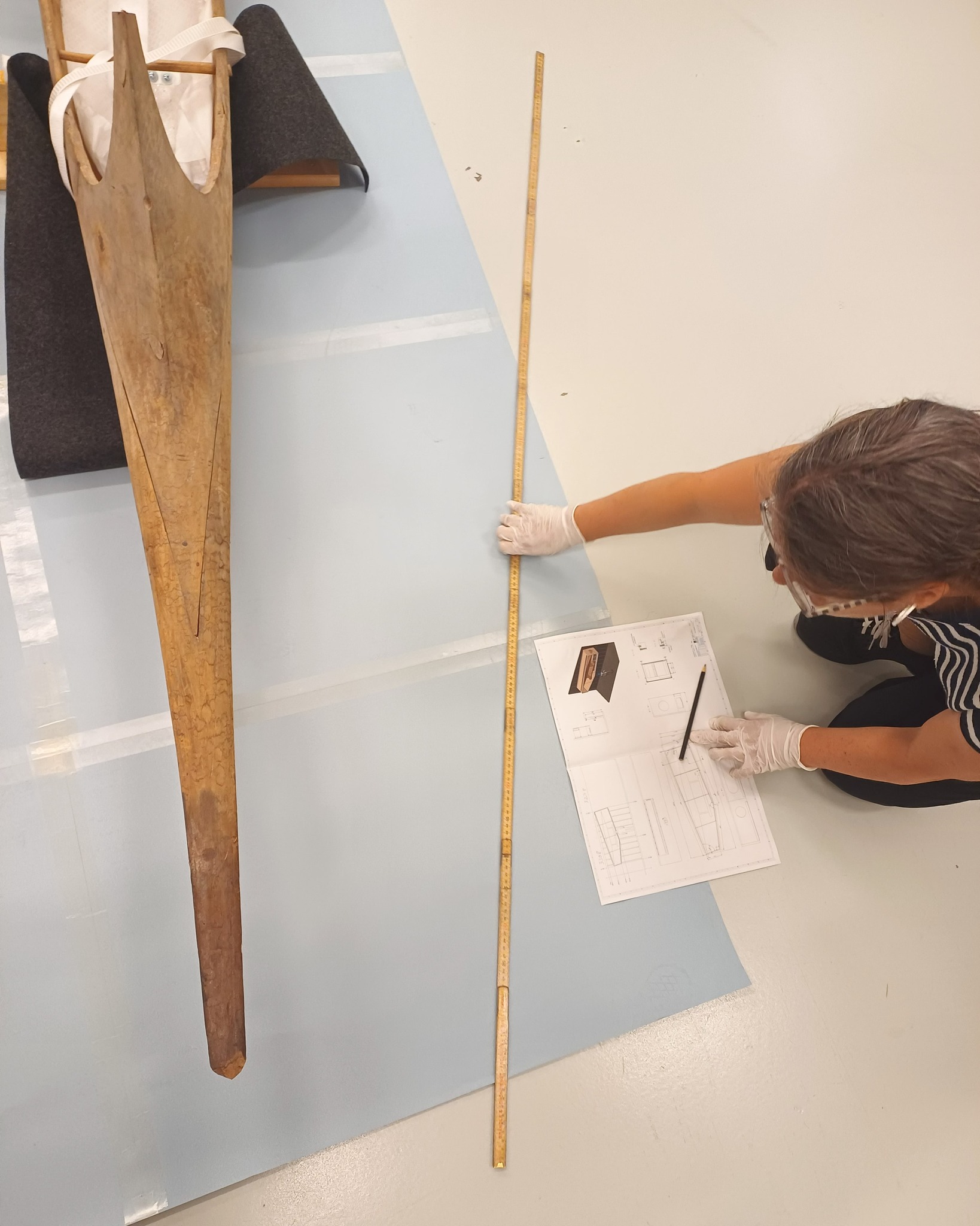 Eine Person mit weißen Handschuhen kniet auf dem Boden und misst mit einem langen Holzlineal ein langes Holzteil auf einer blauen Unterlage. Daneben liegen technische Zeichnungen mit einem Bleistift darauf. (KI-Alternativtext)