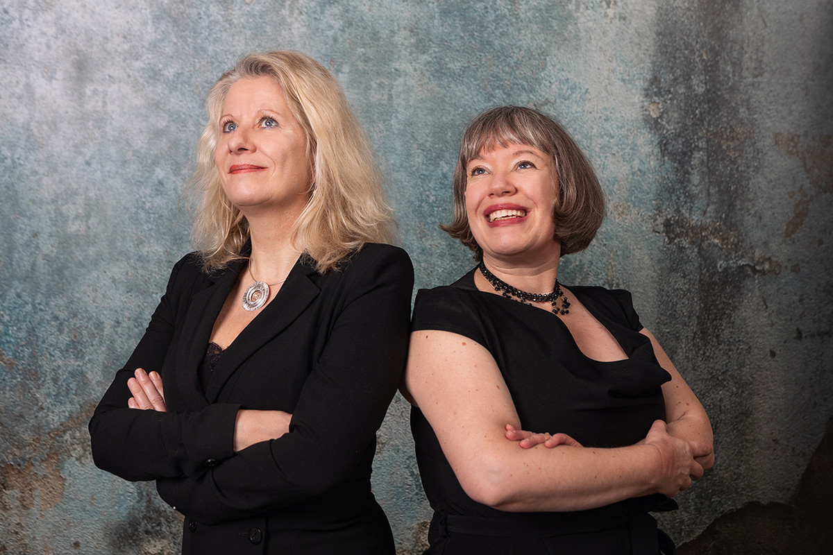 Dieser Alternativtext wurde KI-generiert: Zwei Personen mit hellen Haaren blicken in verschiedene Richtungen. Sie tragen dunkle Kleidung und stehen vor einem strukturierten, hellen Hintergrund.