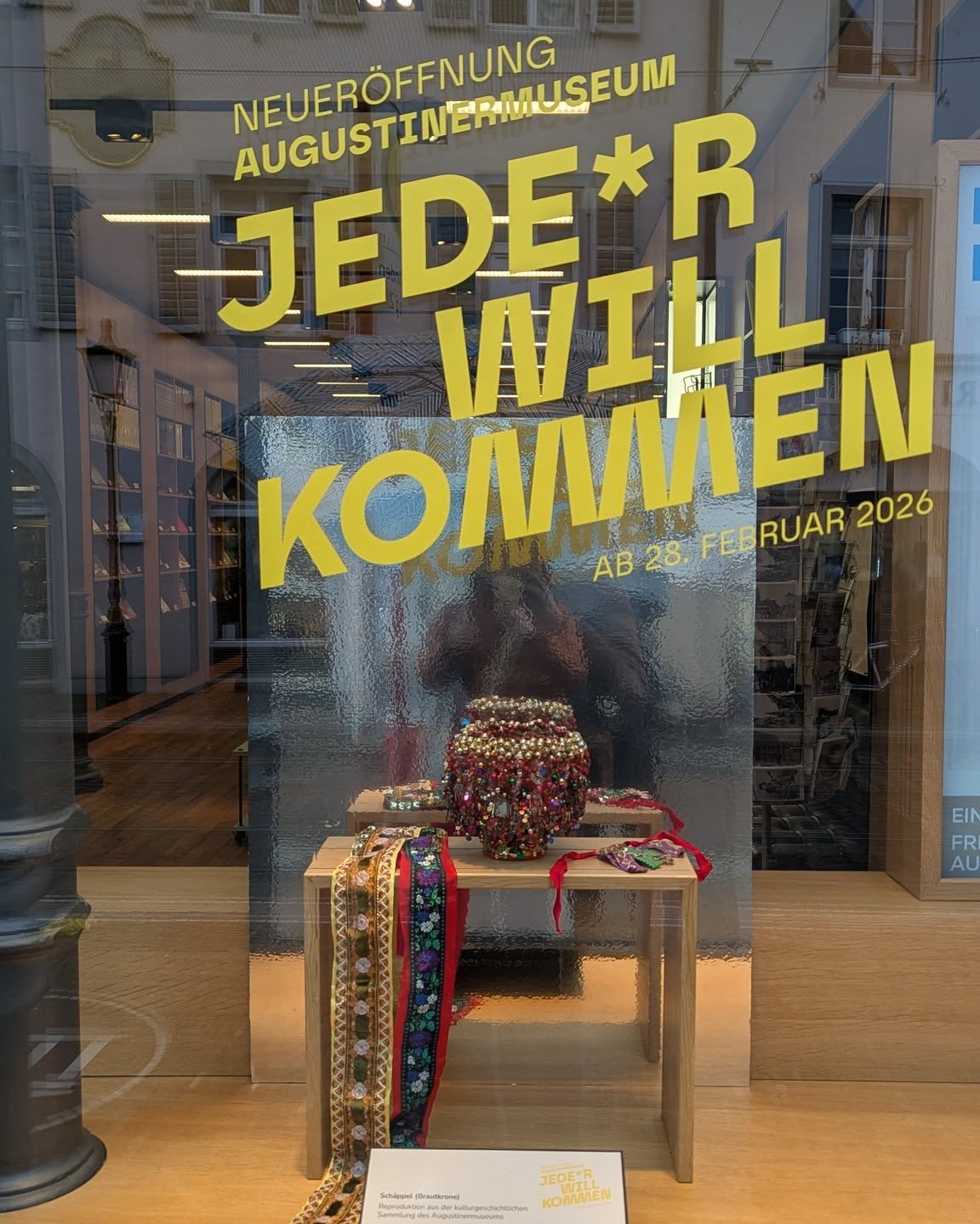 Dieser Alternativtext wurde KI-generiert: Ein Schaufenster mit einer Ankündigung für eine Ausstellung im Februar 2026. Im Schaufenster sind dekorative Objekte und ein farbenfrohes Band zu sehen.