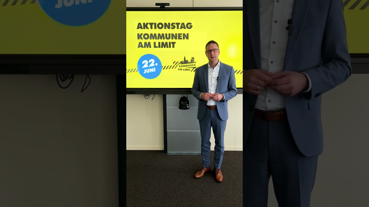 Eine Person in Anzug und Brille steht vor einem Bildschirm mit der Aufschrift „Aktionstag Kommunen am Limit, 22. Juni“. Der Hintergrund ist gelb mit blauen und schwarzen Textelementen. (KI-Alternativtext)