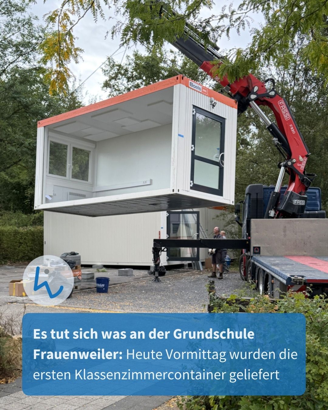 Ein weißer Container mit Fenster und Tür wird von einem roten Kranwagen gehoben, während zwei Personen im Hintergrund stehen. Ein blauer Textkasten am unteren Rand lautet: „Es tut sich was an der Grundschule Frauenweiler: Heute Vormittag wurden die ersten Klassenzimmercontainer geliefert“. (KI-Alternativtext)