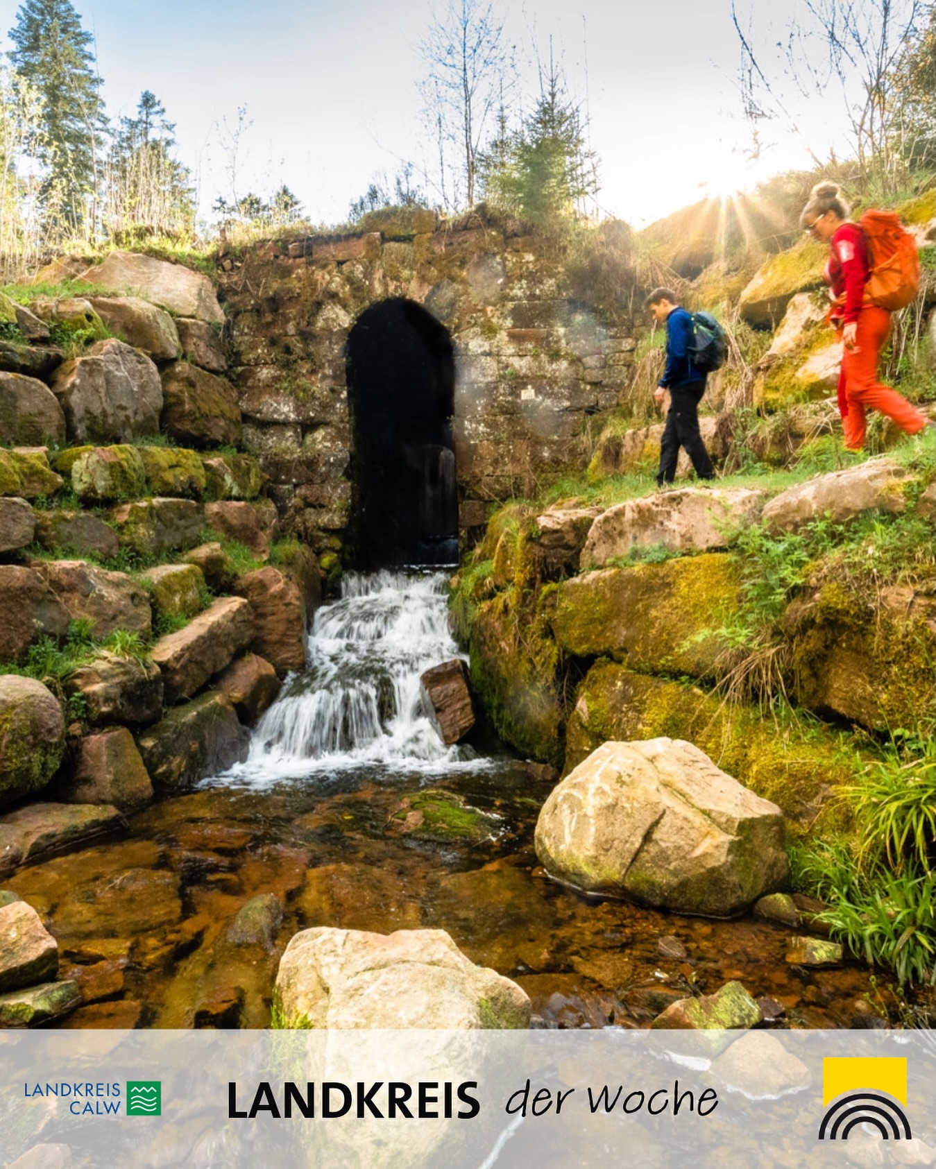 Dieser Alternativtext wurde KI-generiert: Ein Wasserfall fließt unter einem Steintunnel hindurch. Im Hintergrund sind Personen auf einem Pfad in einer hügeligen Landschaft zu sehen.