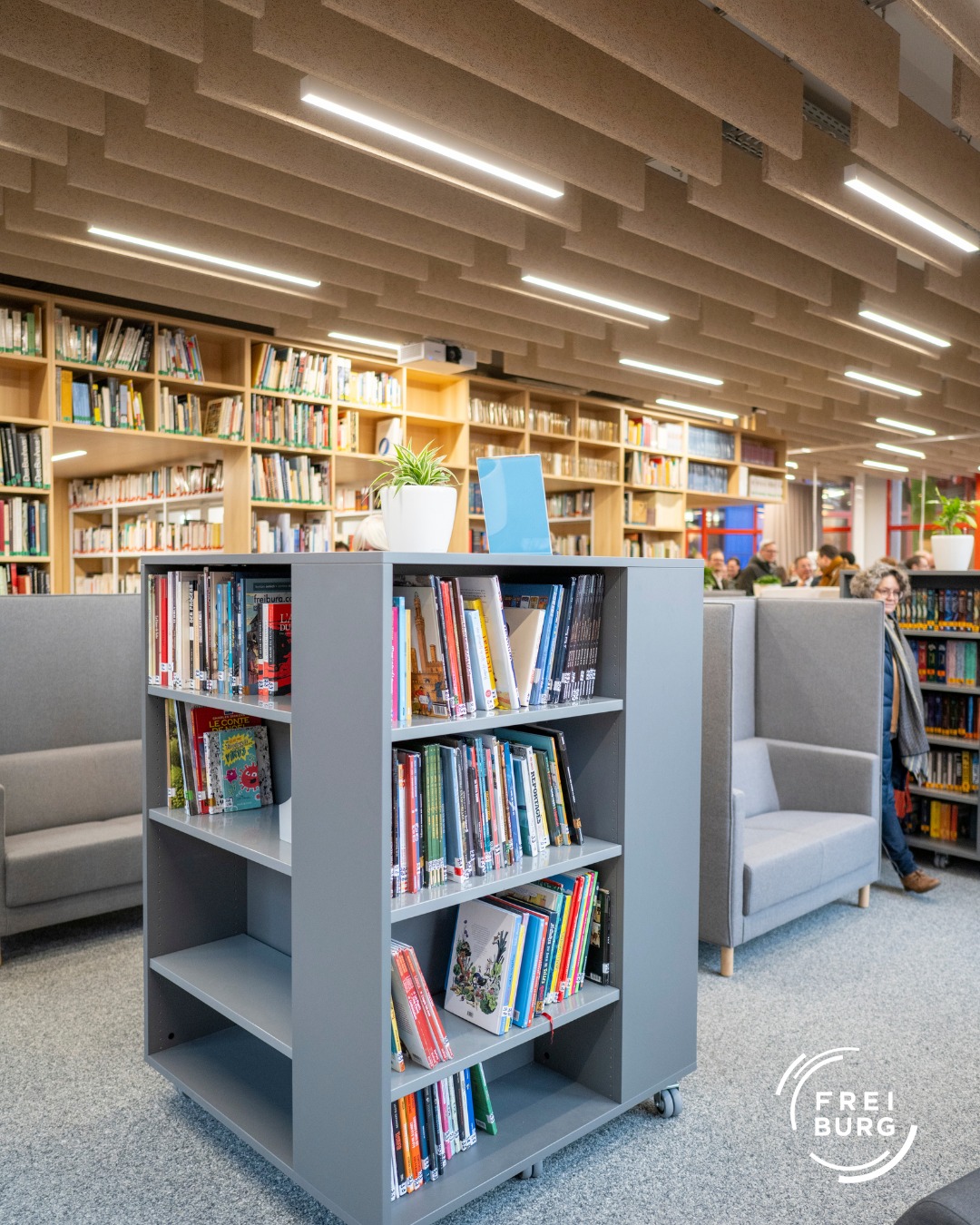 Dieser Alternativtext wurde KI-generiert: Ein Innenraum mit Bücherregalen und Sitzgelegenheiten. Die Decke hat eine interessante Holzstruktur und wird von zahlreichen Leuchten beleuchtet.