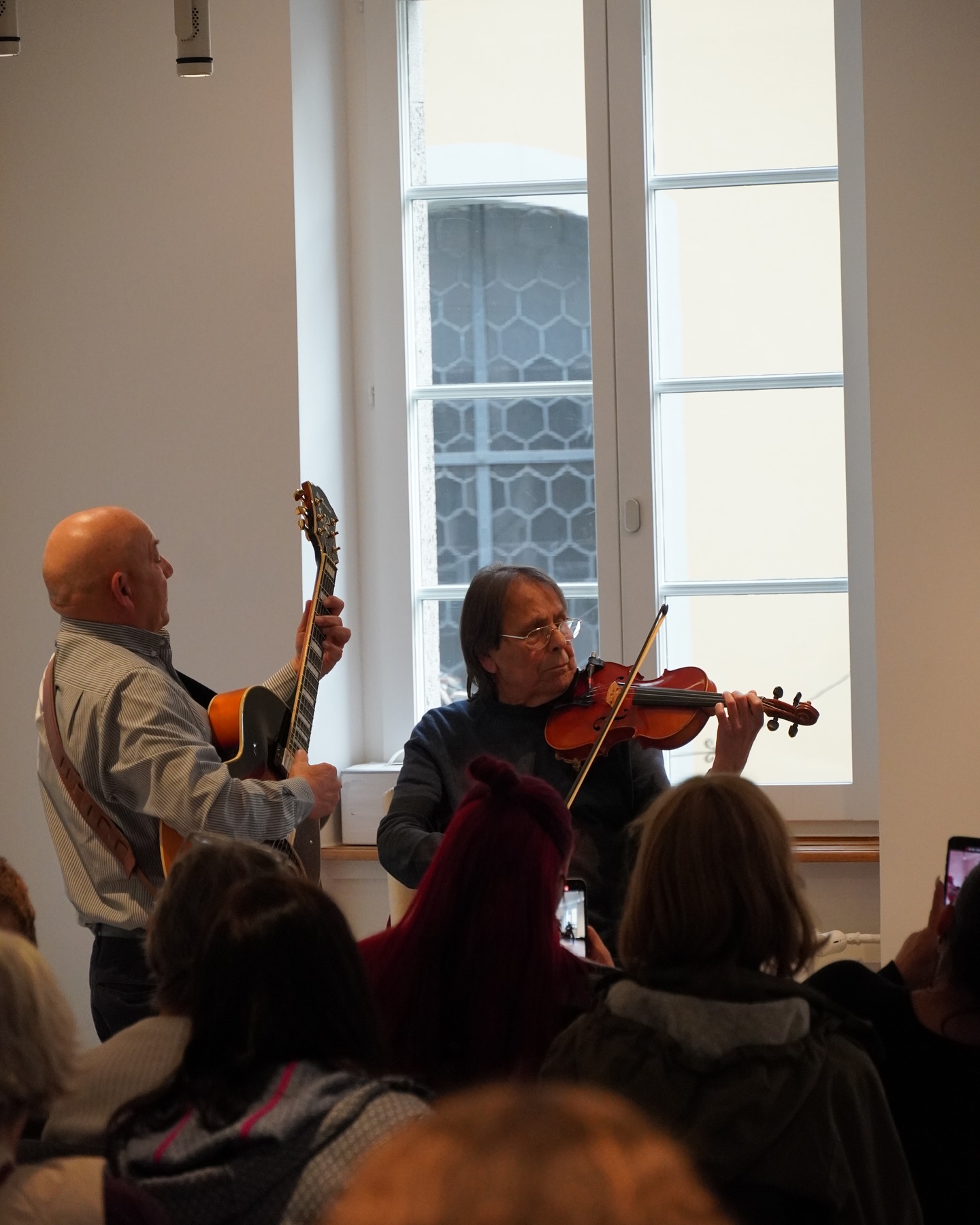 Zwei Musiker spielen vor Publikum: Einer spielt Gitarre, der andere Geige. Das Konzert findet in einem hellen Raum mit Fenster statt.