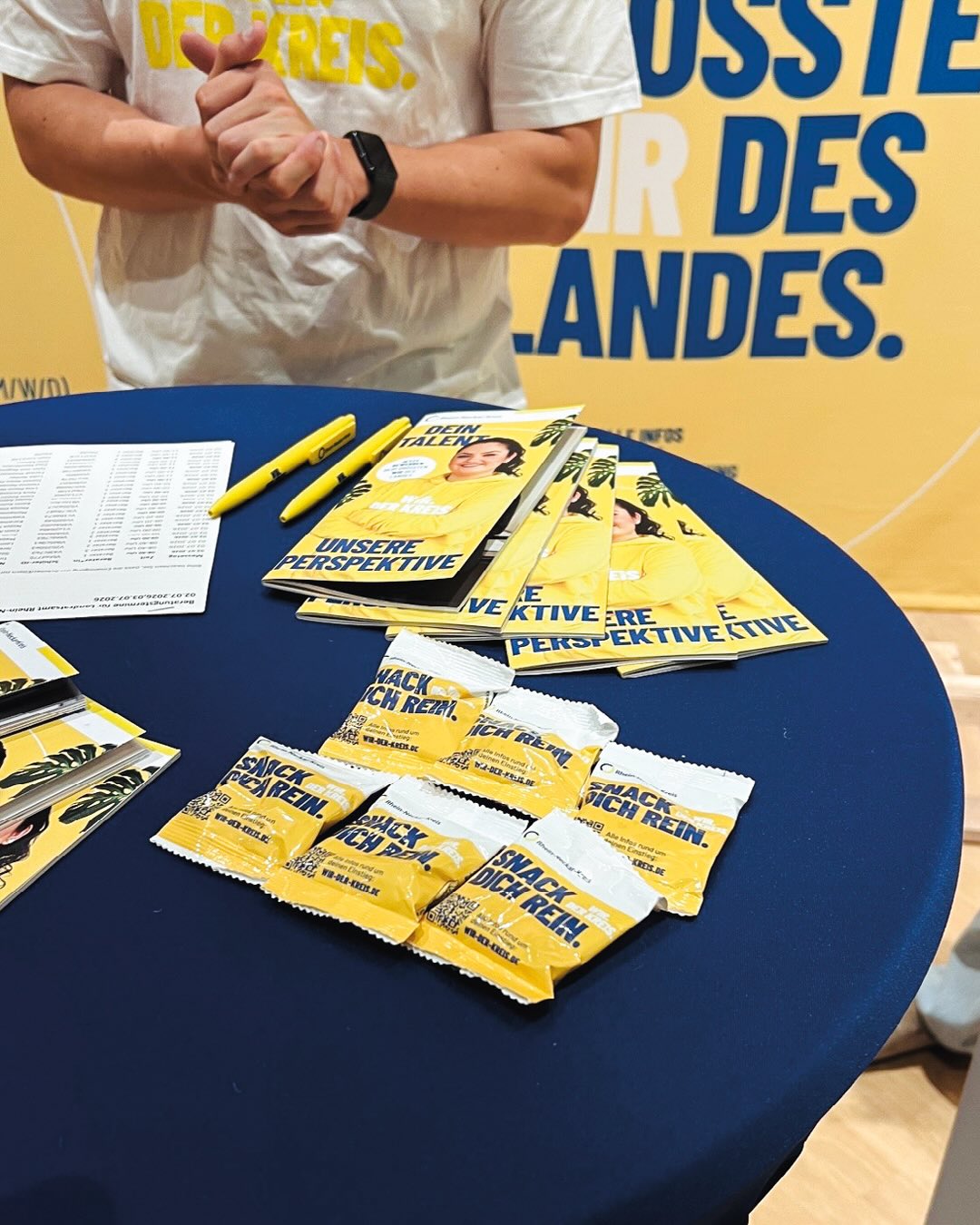 Auf einem blauen Tisch liegen gestapelte gelbe Flyer mit den Aufschriften „Dein Talent“ und „Unsere Perspektive“, gelbe Stifte, ein Listenpapier sowie mehrere kleine Snack-Päckchen mit der Aufschrift „Snack Rein“. Dahinter steht eine Person in einem weißen T-Shirt mit der gelben Aufschrift „DER KREIS.“, die die Hände verschränkt hält, vor einem gelben Hintergrundbanner mit blauen Großbuchstaben. (KI-Alternativtext)