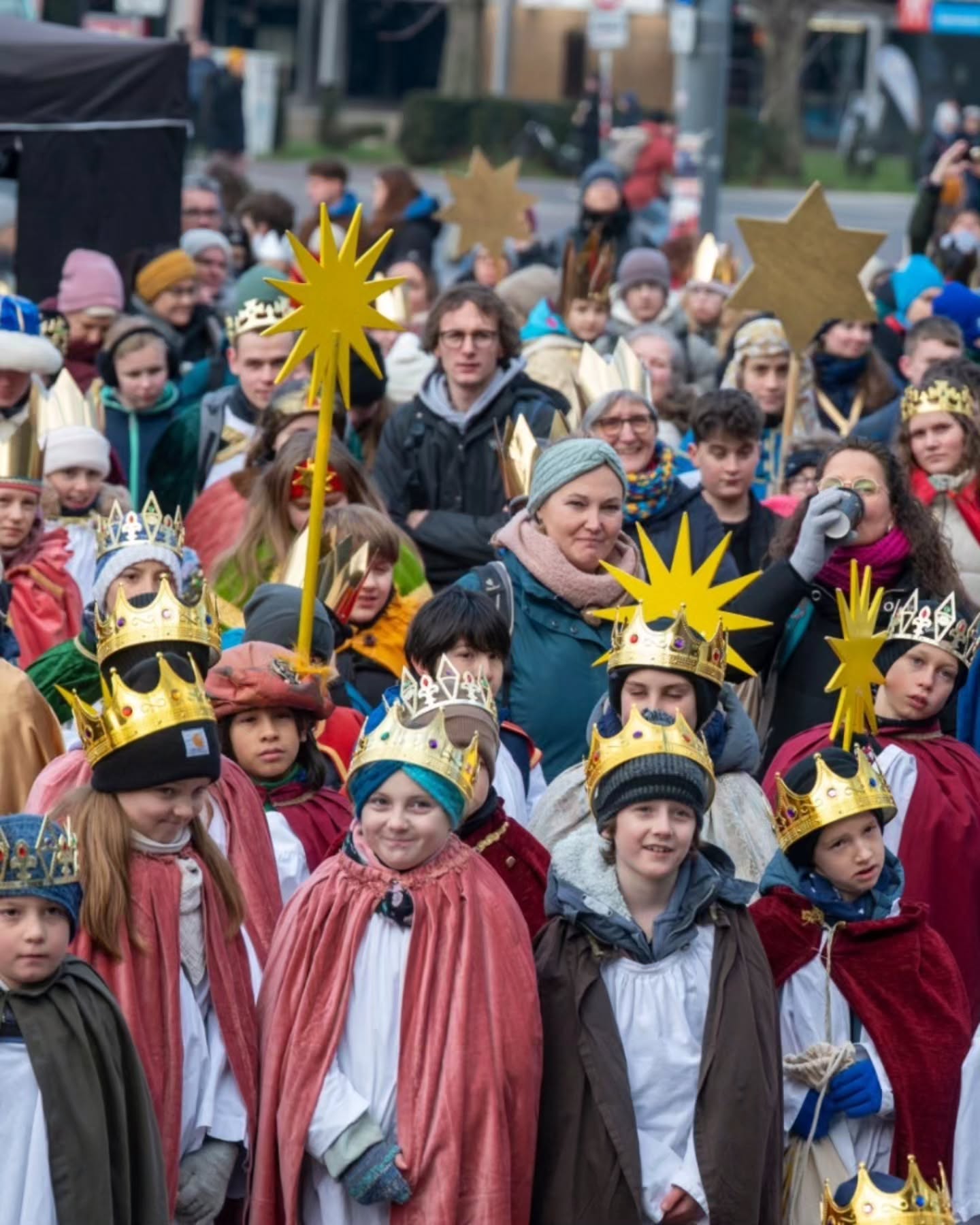 Dieser Alternativtext wurde KI-generiert: Eine Menschengruppe steht oder geht auf einer Straße. Viele Personen tragen Sterne oder Kronen auf dem Kopf und rote Mäntel.