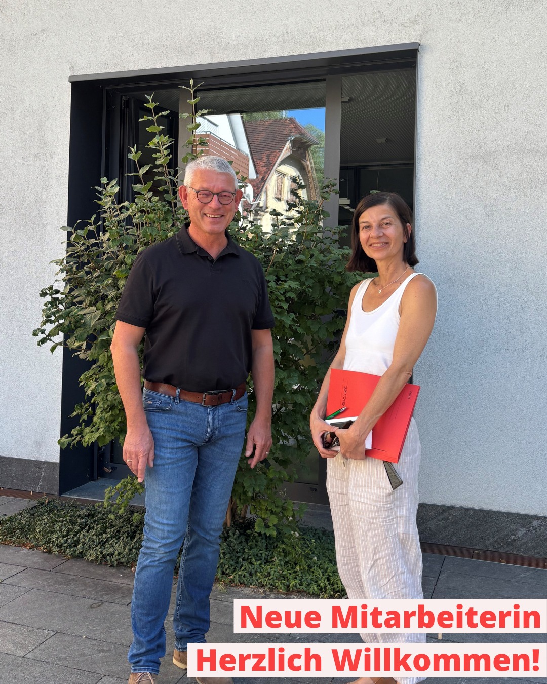 Zwei lächelnde Personen stehen vor einem Gebäude mit Fenster und Büschen. Die Person links trägt ein schwarzes Poloshirt und Jeans, die Person rechts hält einen roten Ordner. Ein Textfeld unten rechts lautet: "Neue Mitarbeiterin Herzlich Willkommen!". (KI-Alternativtext)