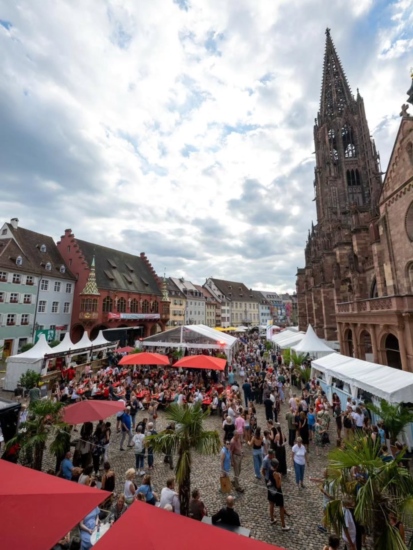 Eine große Menschenmenge bevölkert einen gepflasterten Platz, der mit weißen Zelten und roten Sonnenschirmen für ein Fest oder einen Markt eingerichtet ist. Rechts thront ein hoher gotischer Kirchturm, während links farbenfrohe historische Gebäude stehen. (KI-Alternativtext)