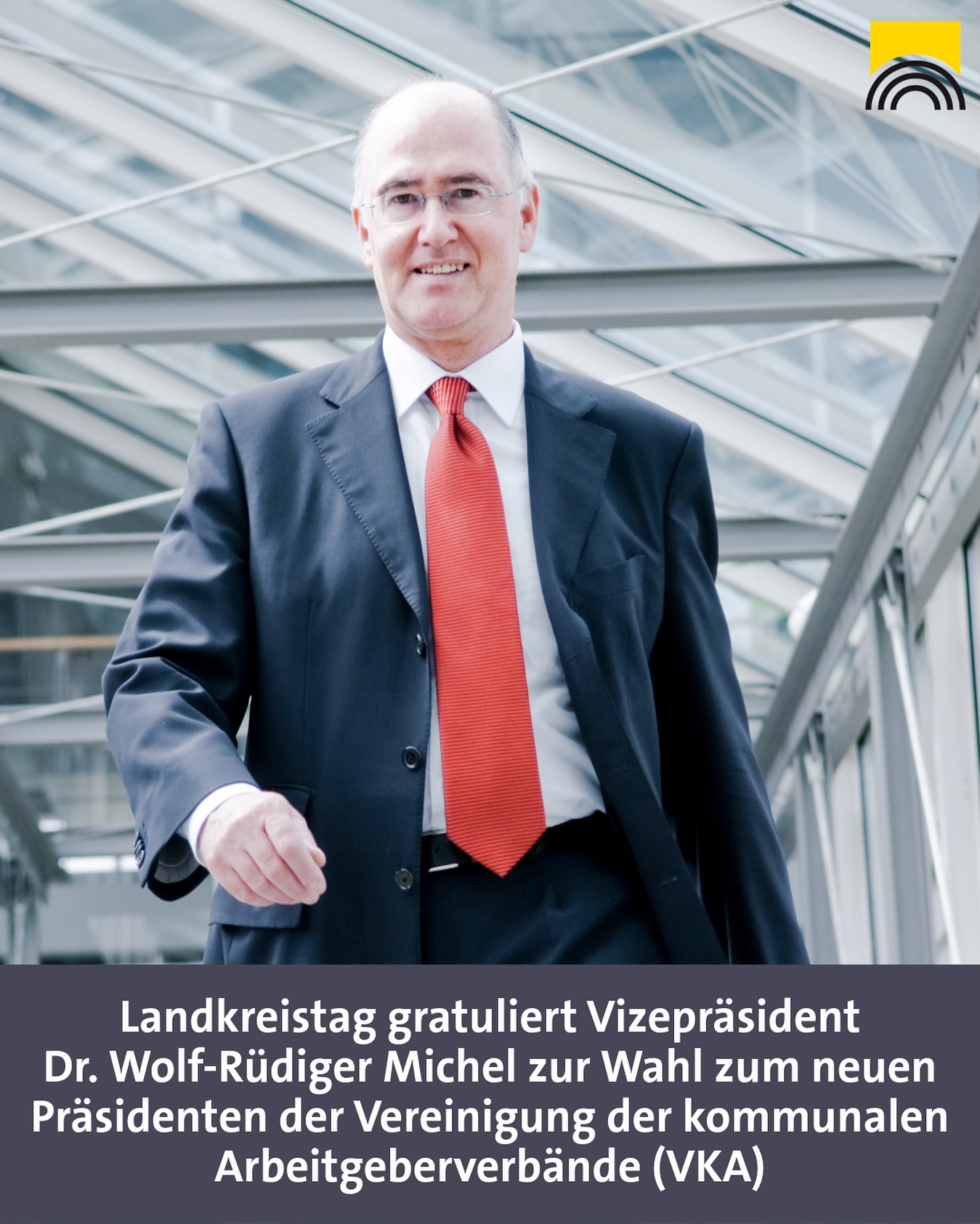 Dieser Alternativtext wurde KI-generiert: Ein Mann in einem Anzug lächelt in einem hellen Raum mit einer Glasfassade. Unter dem Bild befindet sich Text, der eine Gratulation zu einer Wahl ankündigt.