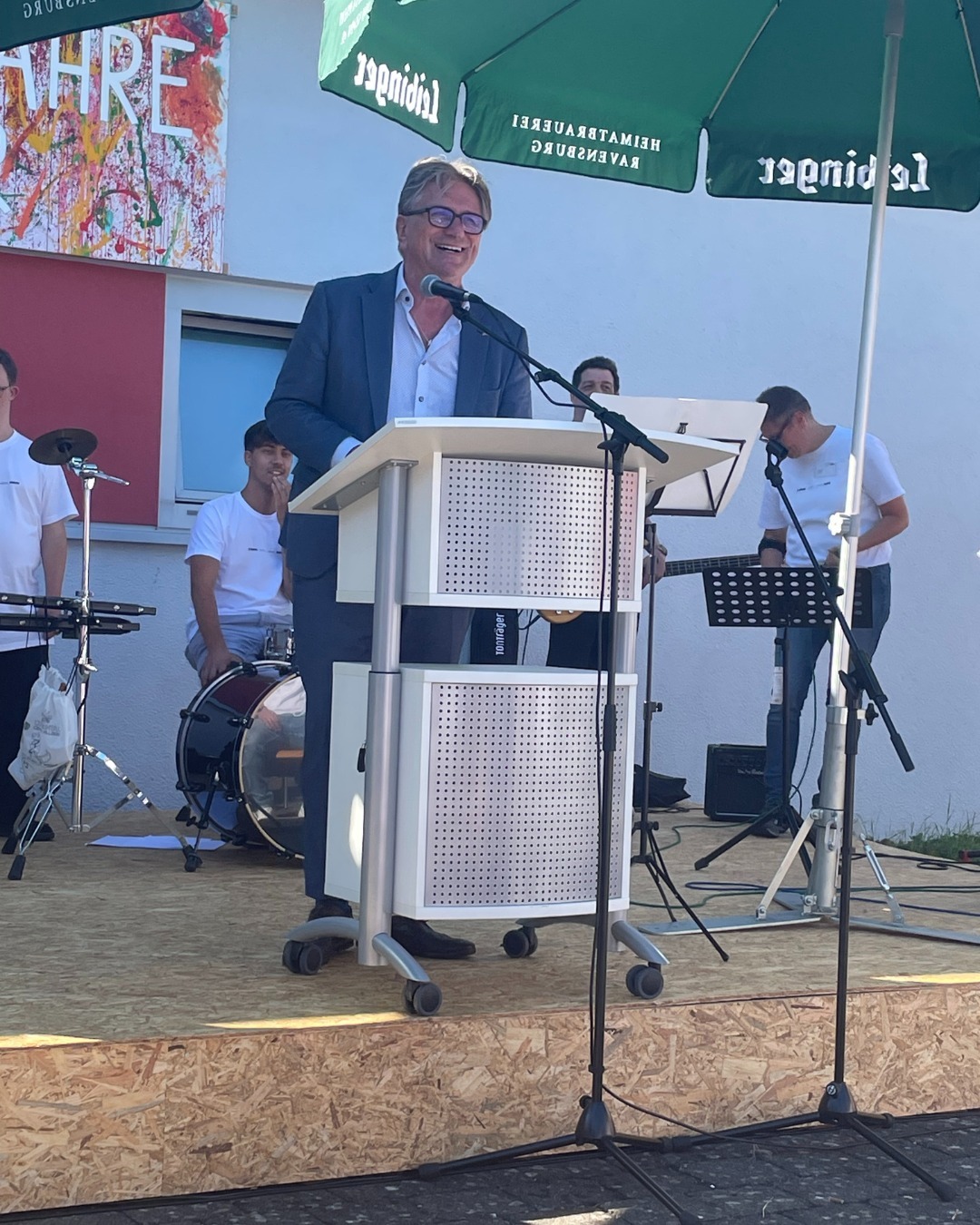 Minister Manne Lucha an einem Rednerpult auf einer Bühne. Im Hintergrund eine Musikband aus jungen Männern.