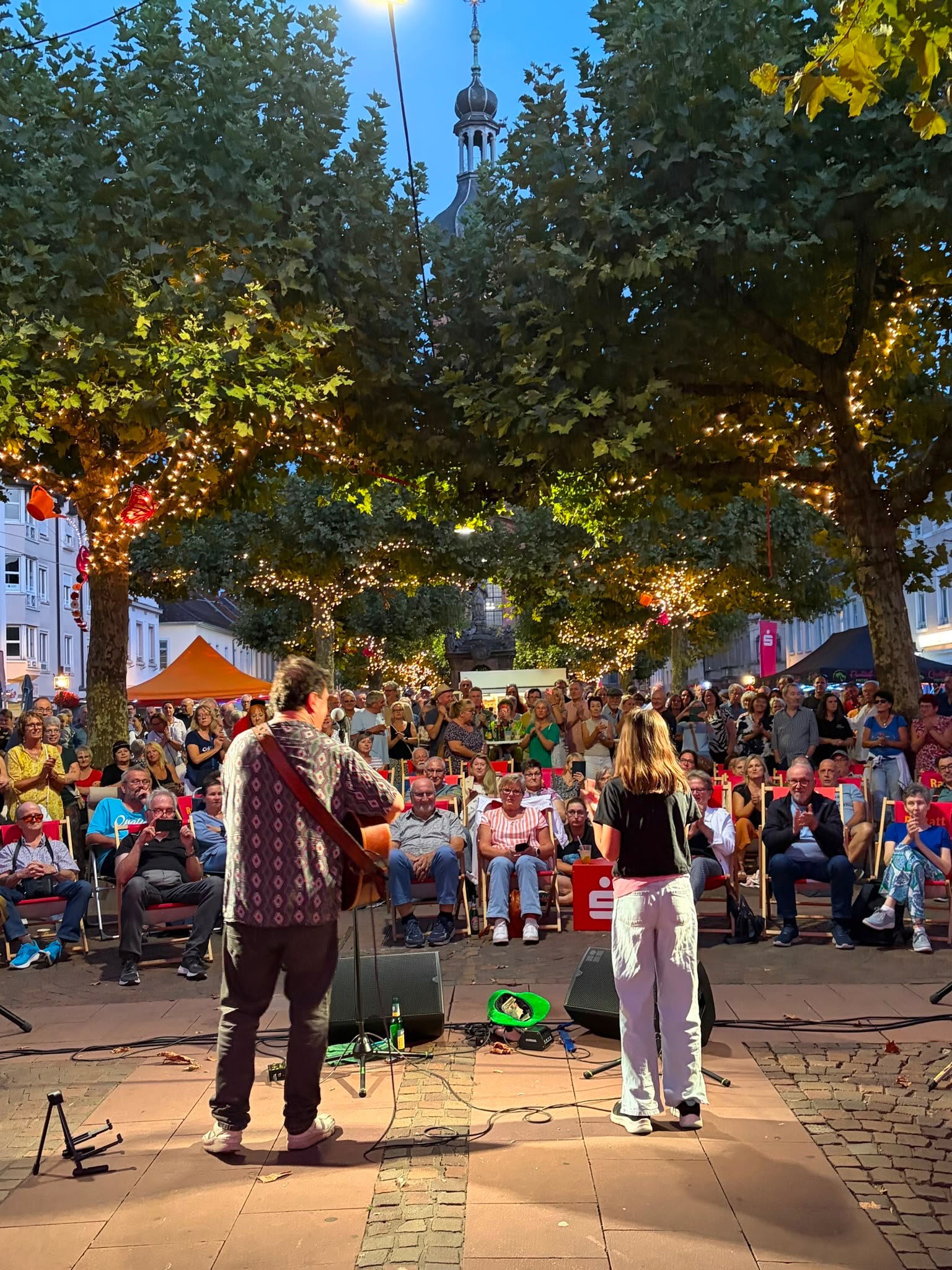 Zwei Personen geben einen musikalischen Auftritt, wobei eine Person Gitarre spielt. Ein großes Publikum sitzt auf roten Stühlen und applaudiert. Im Hintergrund stehen Bäume mit Lichterketten und ein Kirchturm ragt in den Abendhimmel. (KI-Alternativtext)