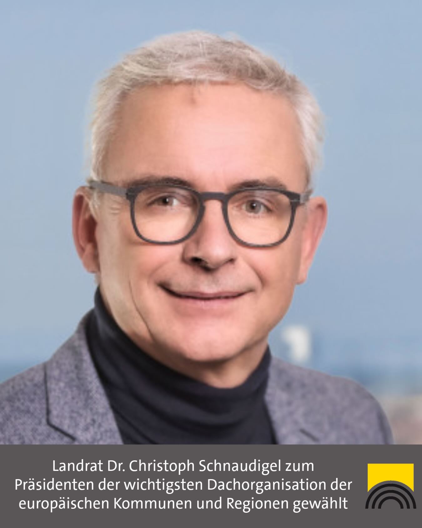 Porträt einer Person mit grauem Haar und Brille, lächelnd, in grauem Sakko und schwarzem Rollkragenpullover vor blauem Hintergrund. Unten Text: „Landrat Dr. Christoph Schnaudigel zum Präsidenten der wichtigsten Dachorganisation der europäischen Kommunen und Regionen gewählt“ sowie ein gelbes Logo mit schwarzen Bögen. (KI-Alternativtext)