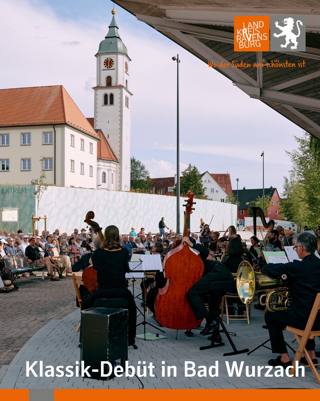 Eine Aussenaufnahme zeigt ein Orchester bei einem Auftritt vor einem Publikum mit einem Turm im Hintergrund.