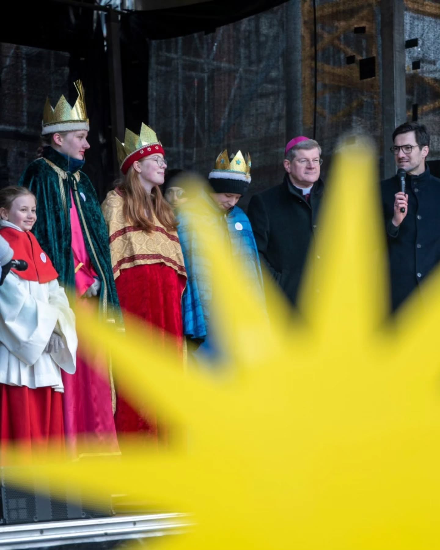 Dieser Alternativtext wurde KI-generiert: Mehrere Personen stehen auf einer Bühne und tragen festliche Kleidung und Kopfbedeckungen. Im Vordergrund ist ein unscharfer, gelber Bereich zu sehen.
