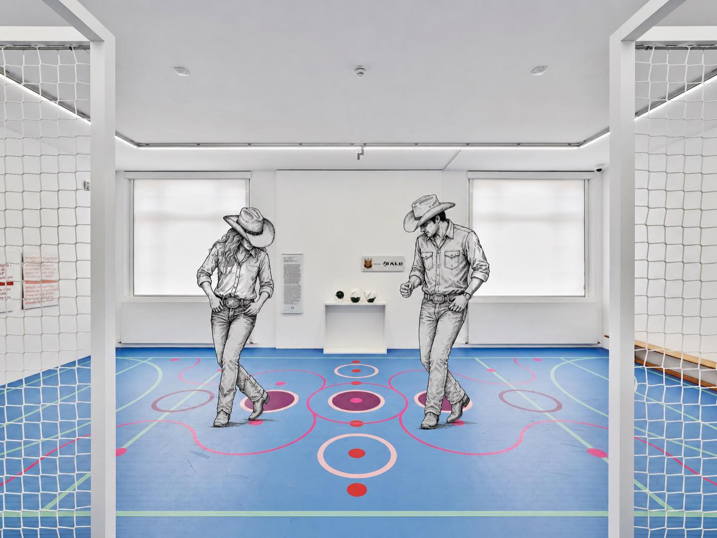 Zwei Personen in Cowboykleidung und -hüten tanzen auf einem blauen Boden mit bunten Kreisen in einem hellen Raum mit weißen Wänden und Fenstern; an den Seiten sind Netzstrukturen sichtbar. (KI-Alternativtext)