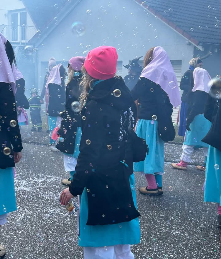 Dieser Alternativtext wurde KI-generiert: Eine Gruppe von Menschen in traditioneller Kleidung steht im Freien. Luftseifen schweben durch die Luft und es ist eine festliche Stimmung erkennbar.