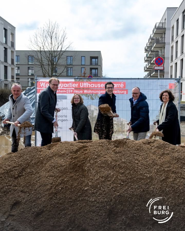 Bild zeigt eine Gruppe von Personen, die an einer symbolischen Grundsteinlegung für ein neues Wohnquartier teilnehmen. Sie halten Spaten in den Händen und stehen vor einem Haufen Erde.