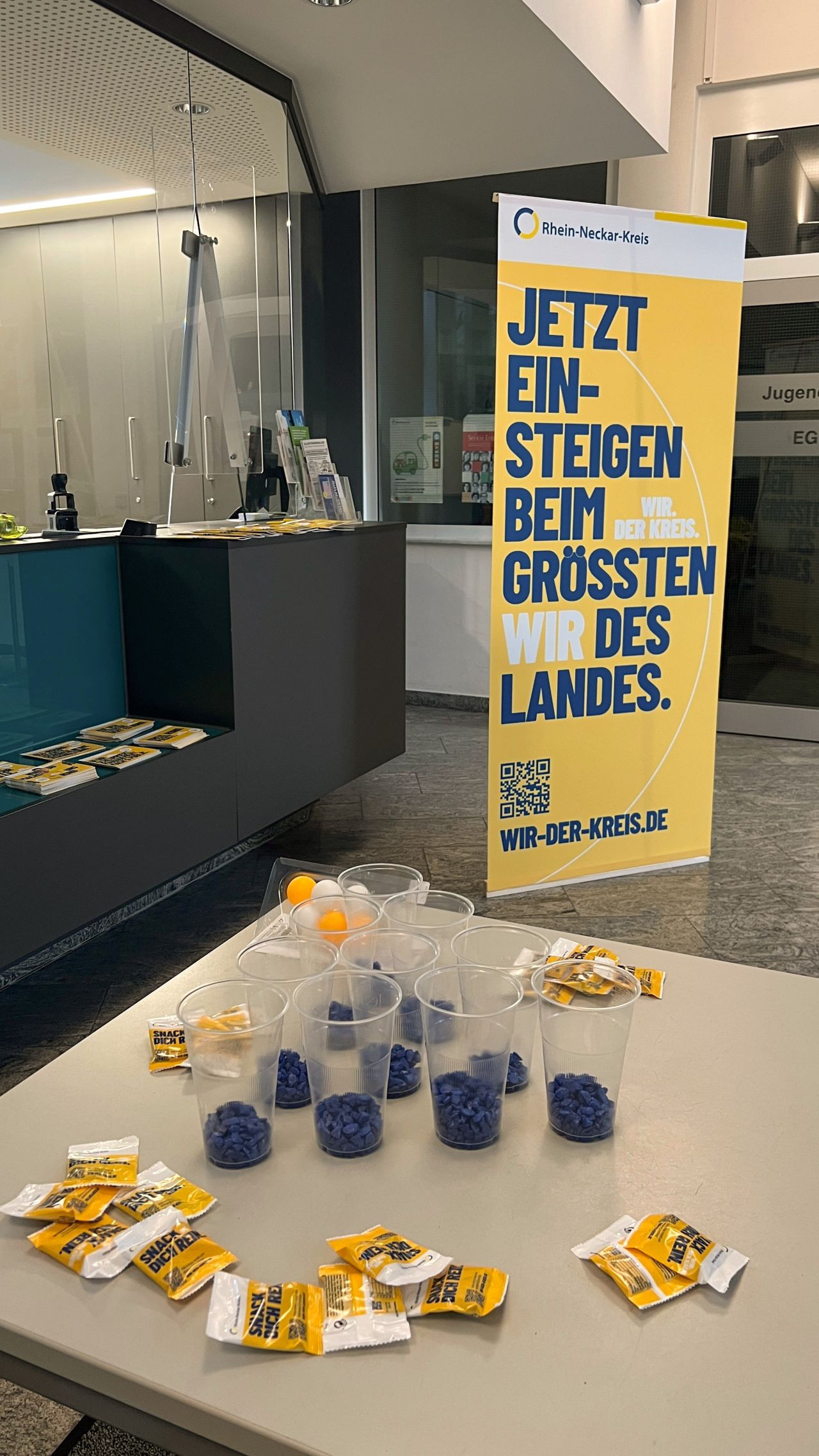 Dieser Alternativtext wurde KI-generiert: Ein Tisch mit kleinen Snacks in verschiedenen Farben und Werbematerialien vor einem Schaufenster. Im Hintergrund ist ein Banner mit Text und einem QR-Code zu sehen.