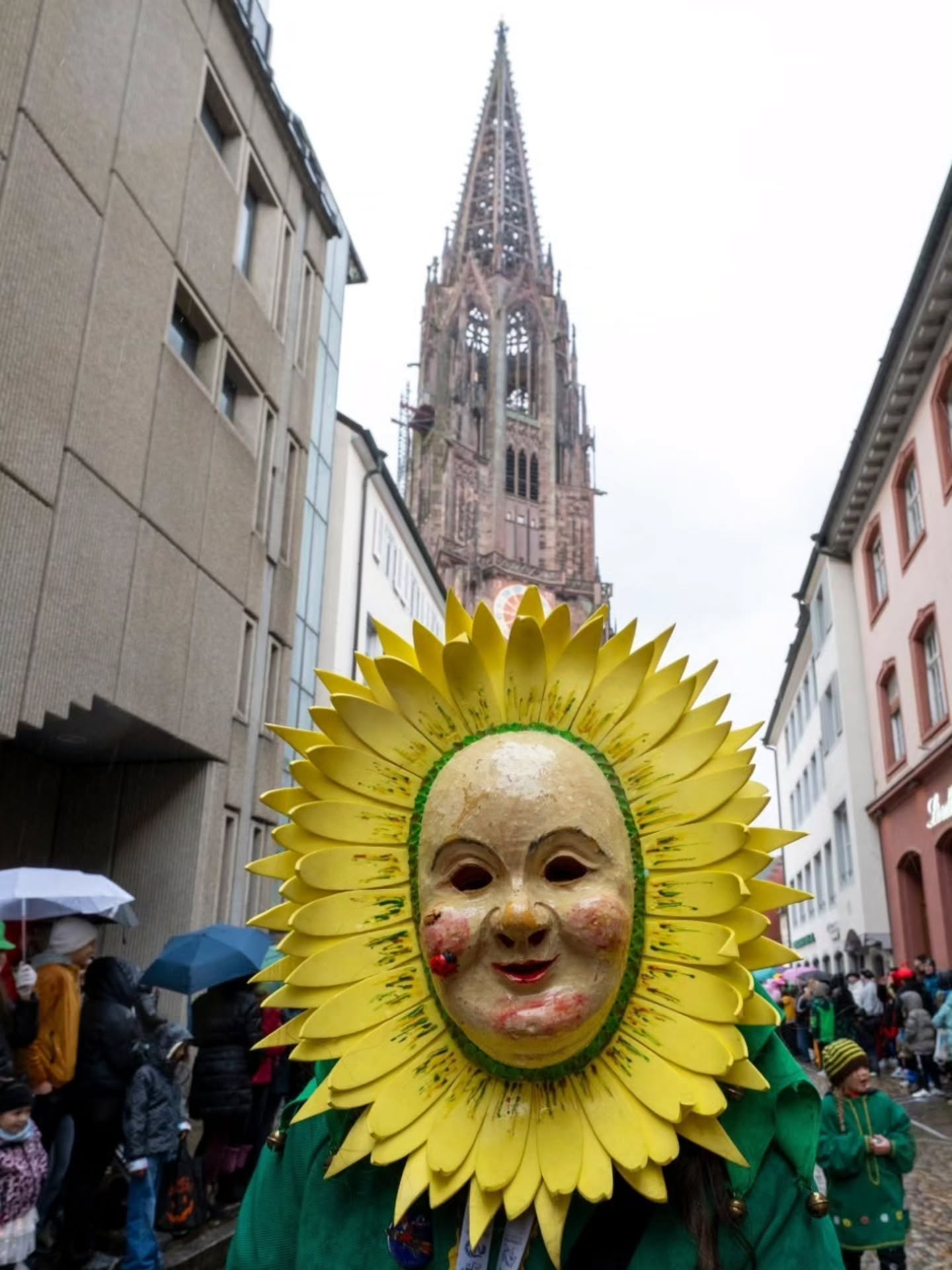 Dieser Alternativtext wurde KI-generiert: Eine Straße ist von hohen Gebäuden gesäumt, am Ende ist ein schlanker Kirchturm zu sehen. Im Vordergrund befindet sich eine Person mit einer großen, sonnenblumenartigen Maske.