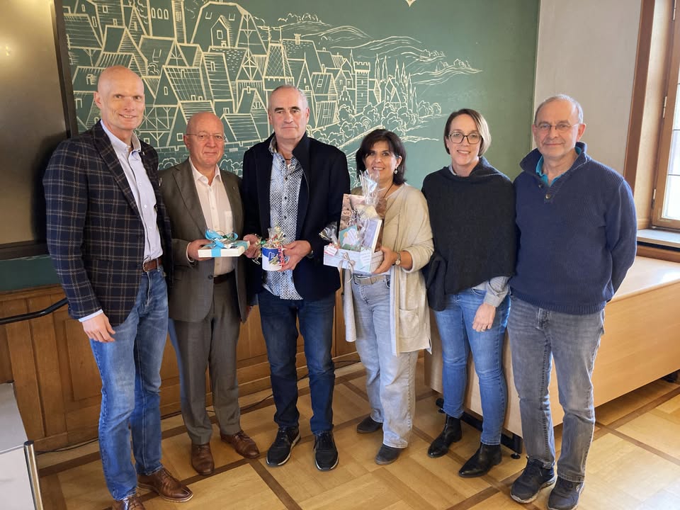 Dieser Alternativtext wurde KI-generiert: Eine Gruppe von Personen steht nebeneinander und hält Geschenke in den Händen. Im Hintergrund befindet sich eine grüne Wand mit einer grafischen Darstellung einer Stadtlandschaft.