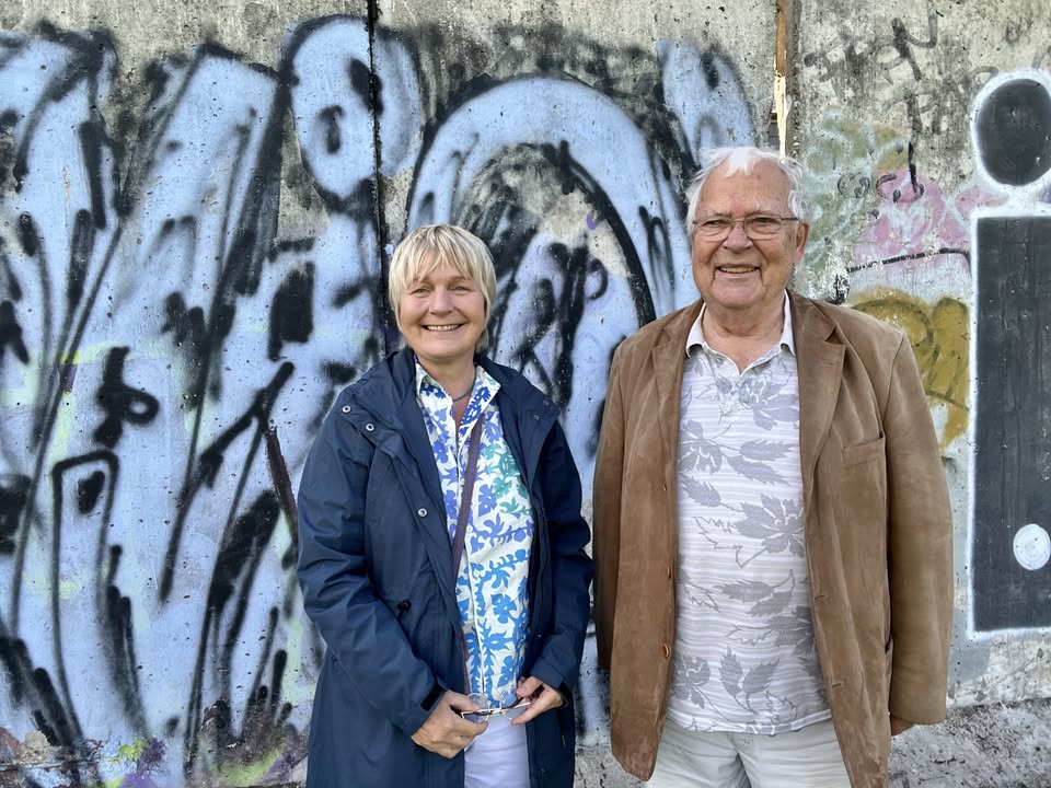 Dieser Alternativtext wurde KI-generiert: Zwei Personen stehen vor einer mit Graffiti beschmierten Wand. Beide lächeln in die Kamera und tragen sommerliche Kleidung.