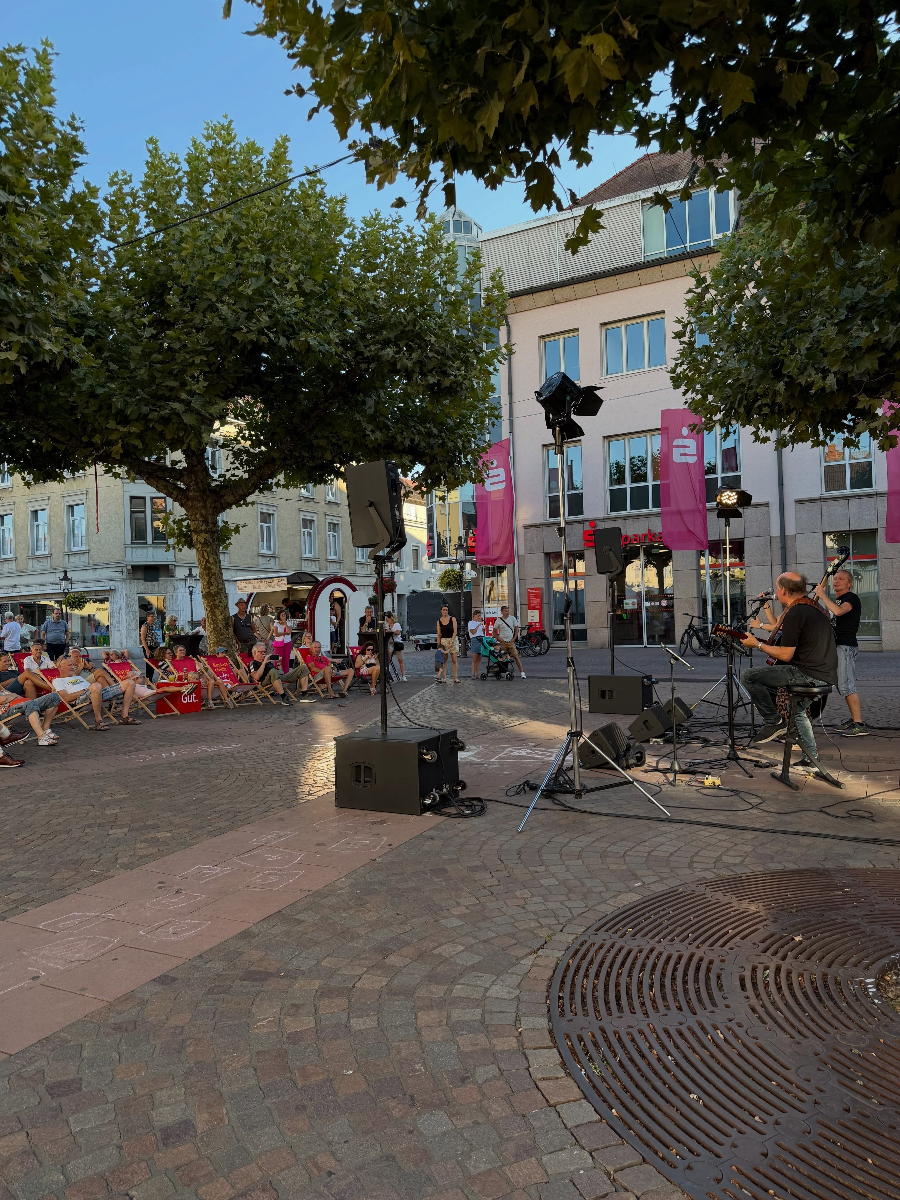 Musiker spielen mit Gitarren und Bass auf einem gepflasterten Platz, umgeben von Lautsprechern und Lichtständern. Links sitzt ein Publikum auf roten Liegestühlen, auf denen „Rastatt chillt“ und „Gut.“ steht, und schaut der Aufführung zu. Im Hintergrund stehen Gebäude, Bäume und ein Food-Truck, während rosa Banner mit einem Logo an einem Gebäude hängen. (KI-Alternativtext)