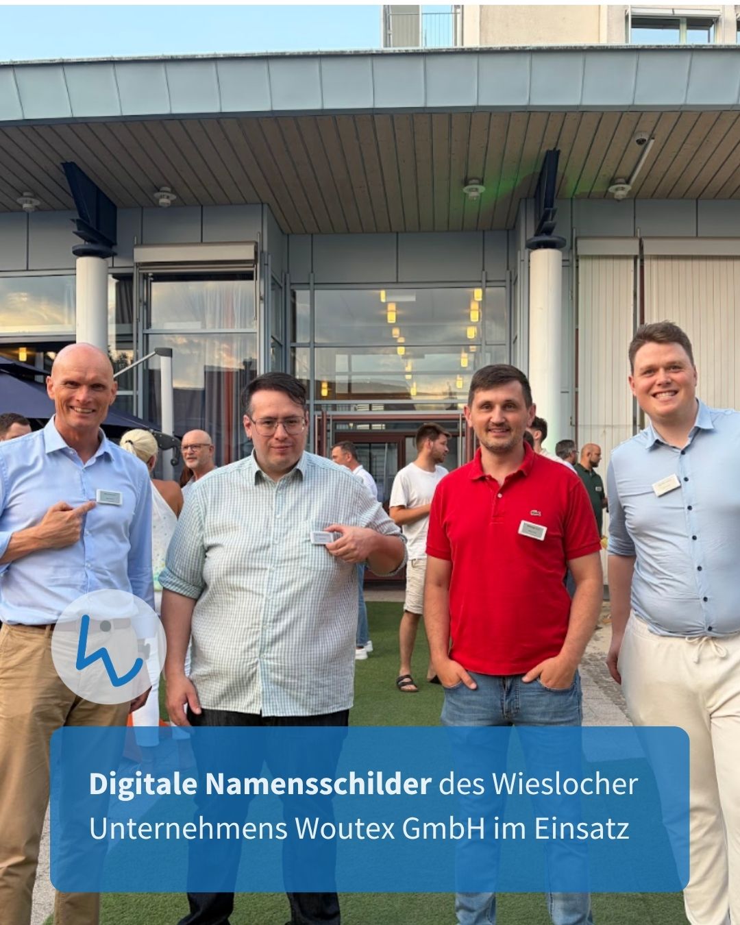 Vier Personen stehen vor einem Gebäudeeingang und präsentieren digitale Namensschilder, wobei zwei auf ihre Brust zeigen und eine ein Schild in der Hand hält. Ein blauer Textbalken unten lautet: "Digitale Namensschilder des Wieslocher Unternehmens Woutex GmbH im Einsatz". (KI-Alternativtext)