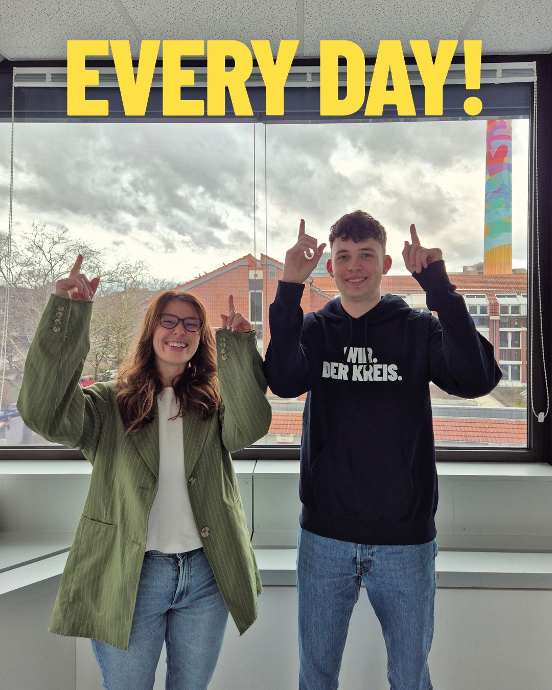 Dieser Alternativtext wurde KI-generiert: Zwei Personen stehen vor einem Fenster mit Blick auf eine Stadtlandschaft und zeigen mit erhobenen Fingern nach oben. Der Hintergrund ist von einem bewölkten Himmel und Gebäuden geprägt.