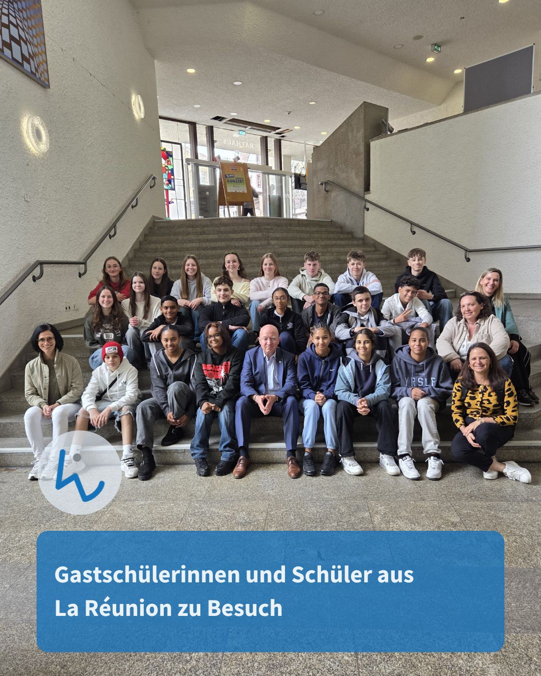 Dieser Alternativtext wurde KI-generiert: Eine Gruppe von Personen sitzt und steht auf einer Treppe in einem Innenraum. Im Hintergrund sind weitere Treppen und Türen zu sehen.