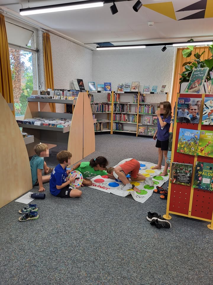 Eine Gruppe von Kindern spielt das Brettspiel Twister auf dem Teppichboden einer Bibliothek. Mehrere Kinder sind auf der bunten Matte verteilt, während eines den Spinner hält und ein weiteres Kind neben einem Bücherregal steht. Im Hintergrund sind weitere Bücherregale und ein roter Buchständer zu sehen. (KI-Alternativtext)