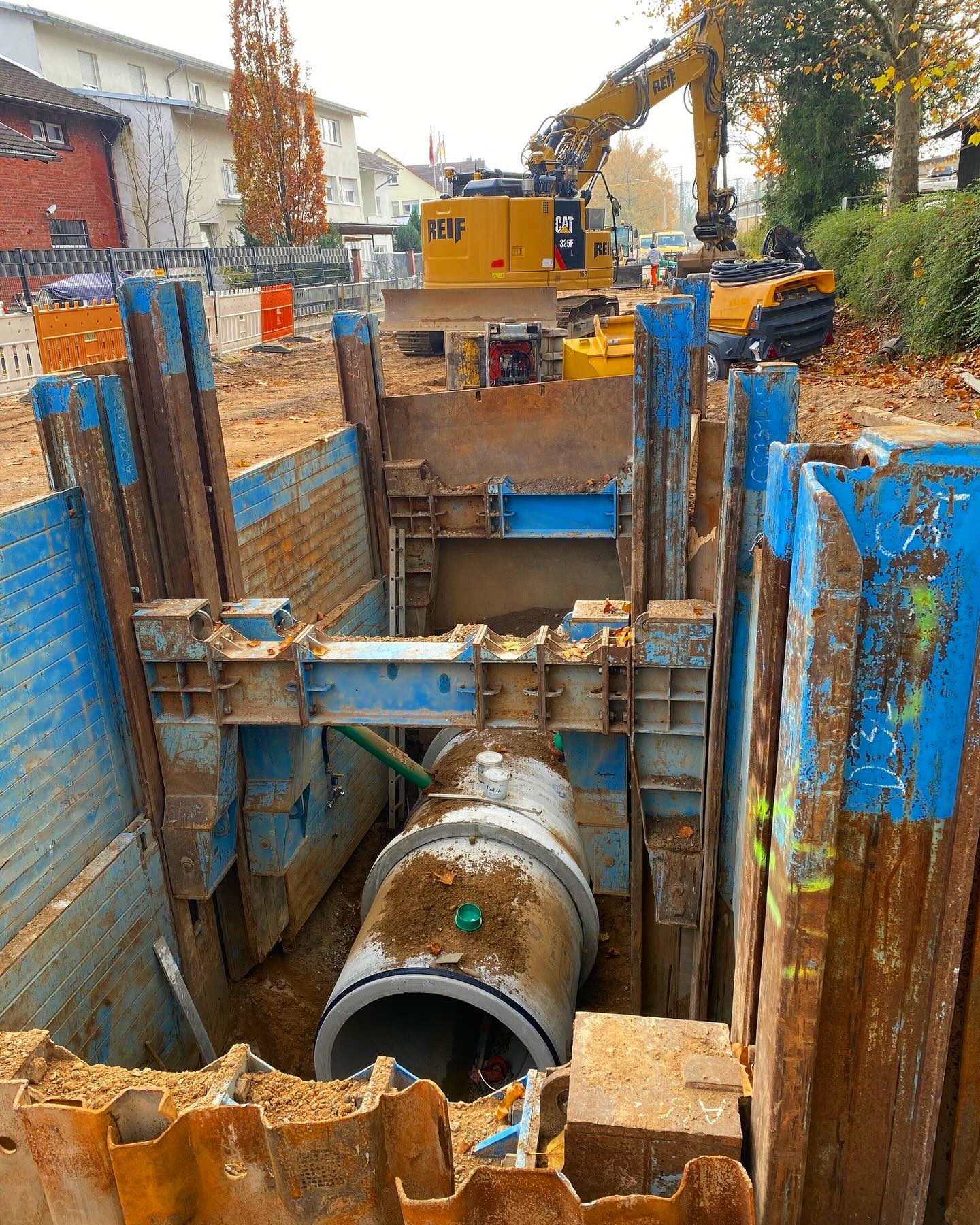 Dieser Alternativtext wurde KI-generiert: Eine Baustelle mit einem großen gelben Bagger und einer Baugrube. Ein Rohr wird in die Grube eingelassen, die Wände sind mit blauen Brettern gesichert.