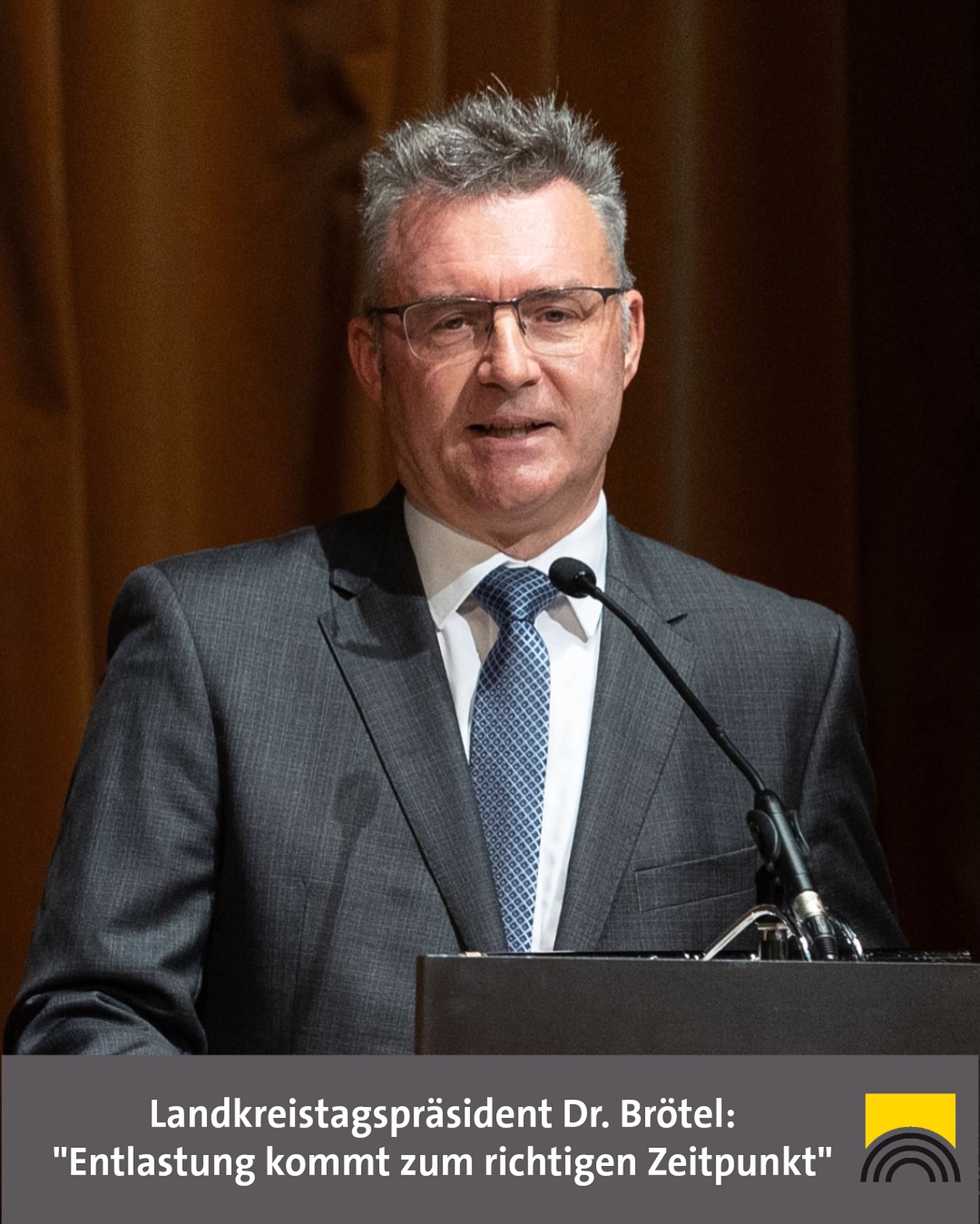 Dieser Alternativtext wurde KI-generiert: Ein Mann mit Brille spricht in ein Mikrofon vor einem Pult. Das Bild ist farbig und zeigt eine Person mit dunklem Anzug und Hemd.