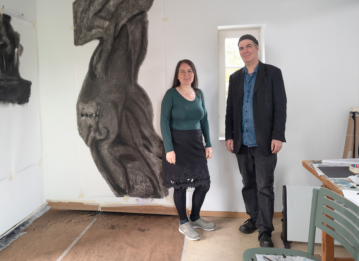 Dieser Alternativtext wurde KI-generiert: Zwei Personen stehen in einem hellen Raum vor einer großen, graustufigen Zeichnung. Im Hintergrund ist ein Fenster und ein Arbeitsbereich zu sehen.