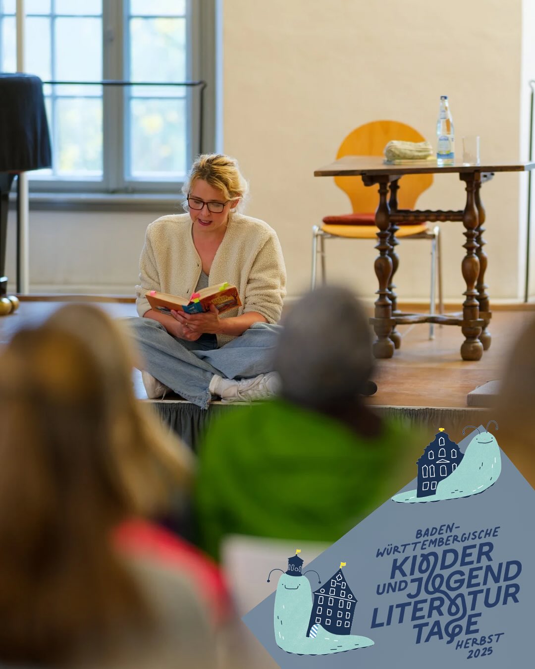 Dieser Alternativtext wurde KI-generiert: Eine Person sitzt auf einem Teppich vor einem Fenster und liest ein Buch. Im Vordergrund befindet sich ein farbenfrohes Plakat mit Illustrationen und Text.