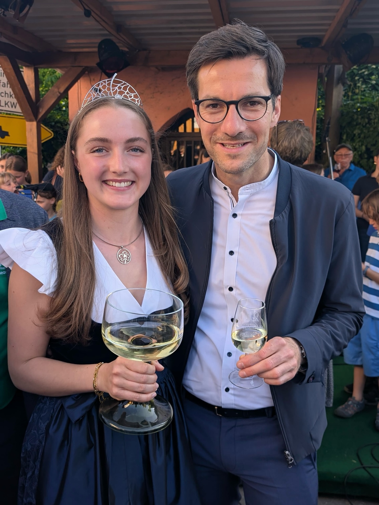Dieser Alternativtext wurde KI-generiert: Zwei Personen stehen lächelnd zusammen und halten Weingläser in der Hand. Im Hintergrund sind weitere Menschen und eine Gartenlandschaft zu sehen.