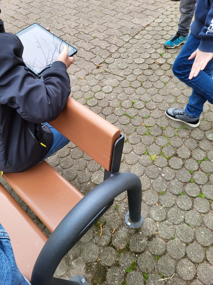 Dieser Alternativtext wurde KI-generiert: Zwei Personen sitzen oder stehen auf einem gepflasterten Weg in der Nähe einer Bank. Eine Person benutzt ein Tablet-Gerät.