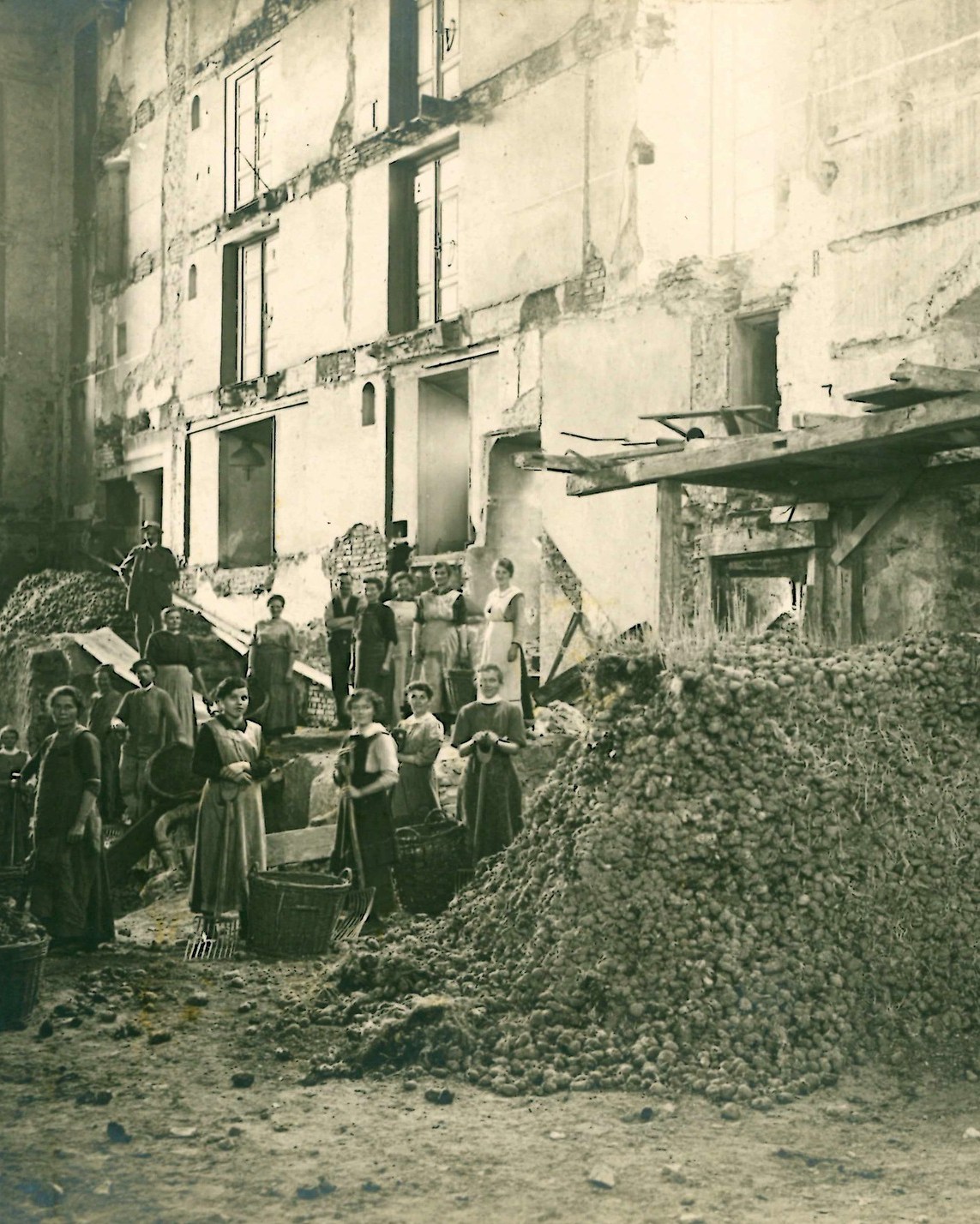 Dieser Alternativtext wurde KI-generiert: Schwarz-weißes historisches Foto einer Gebäudefassade mit beschädigter Struktur. Eine Gruppe von Menschen steht und arbeitet vor einem großen Haufen von etwas, das wie Kartoffeln aussieht.