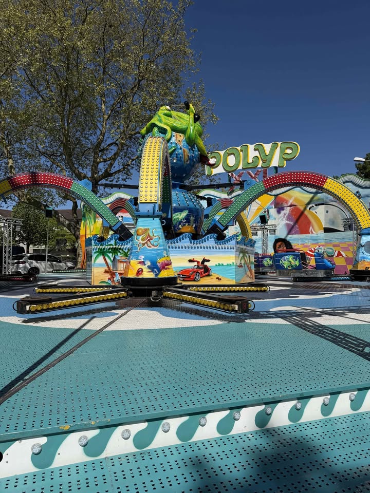 Dieser Alternativtext wurde KI-generiert: Ein farbenfrohes Fahrgeschäft steht auf einem gepflasterten Platz. Im Hintergrund sind Bäume und ein blauer Himmel zu sehen.