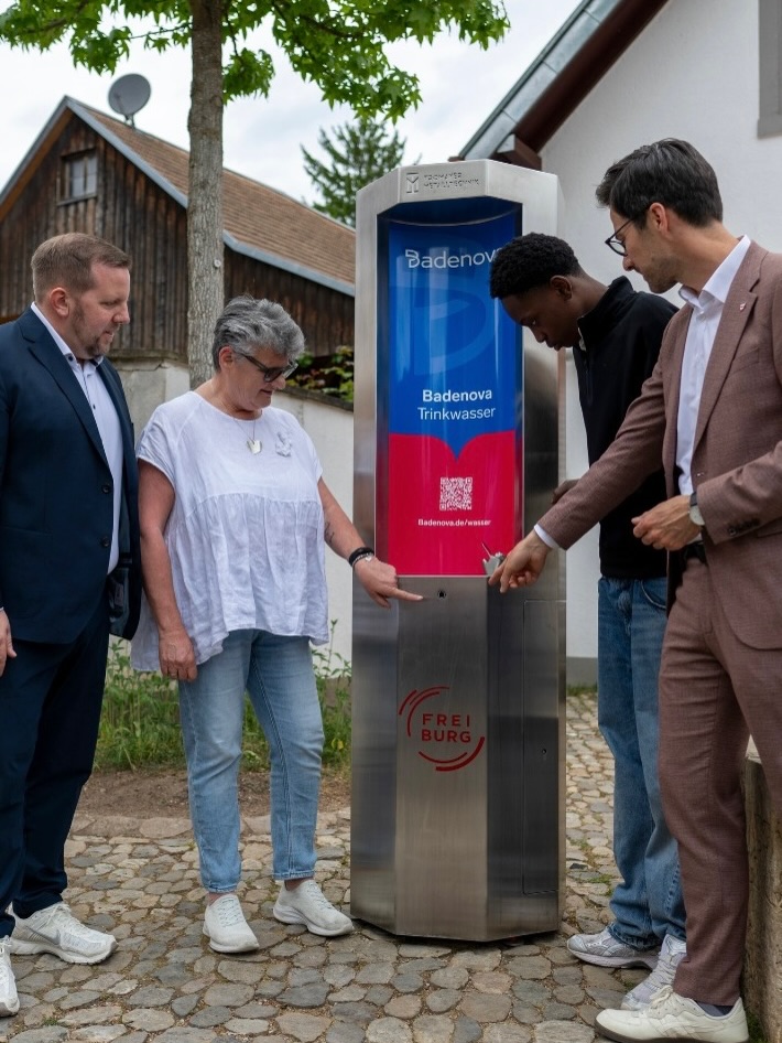 Dieser Alternativtext wurde KI-generiert: Eine farbige Aufnahme zeigt Personen vor einem Gerät zur Ausgabe von Trinkwasser. Das Gerät hat ein Display mit einem QR-Code und steht auf einem gepflasterten Untergrund.